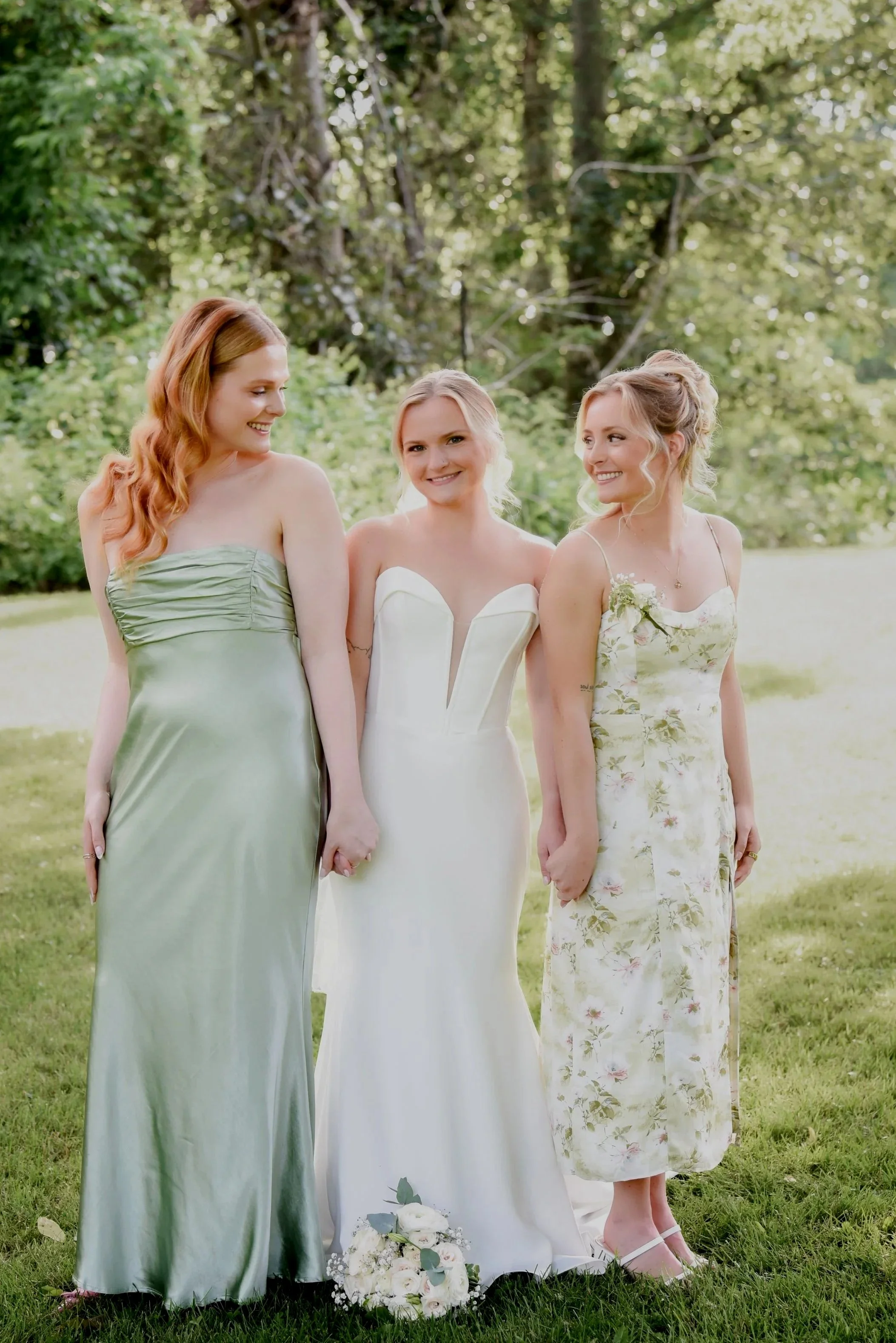 Three women standing outside on grass at a wedding, holding hands, wearing dresses, with a bouquet on the ground near their feet. The woman in the middle is in a white wedding dress, flanked by two women in dresses, one in green and the other in a fl