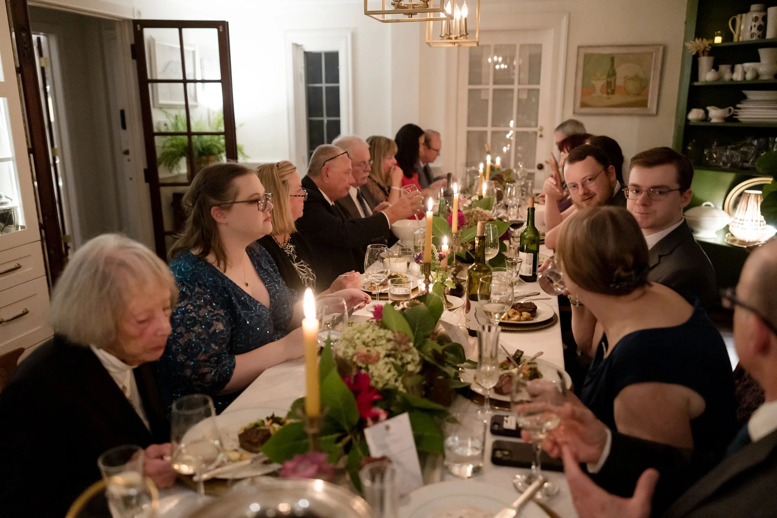 People gathered around a long dinner table, celebrating with lit candles, wine bottles, and festive decorations in a warmly lit dining room.