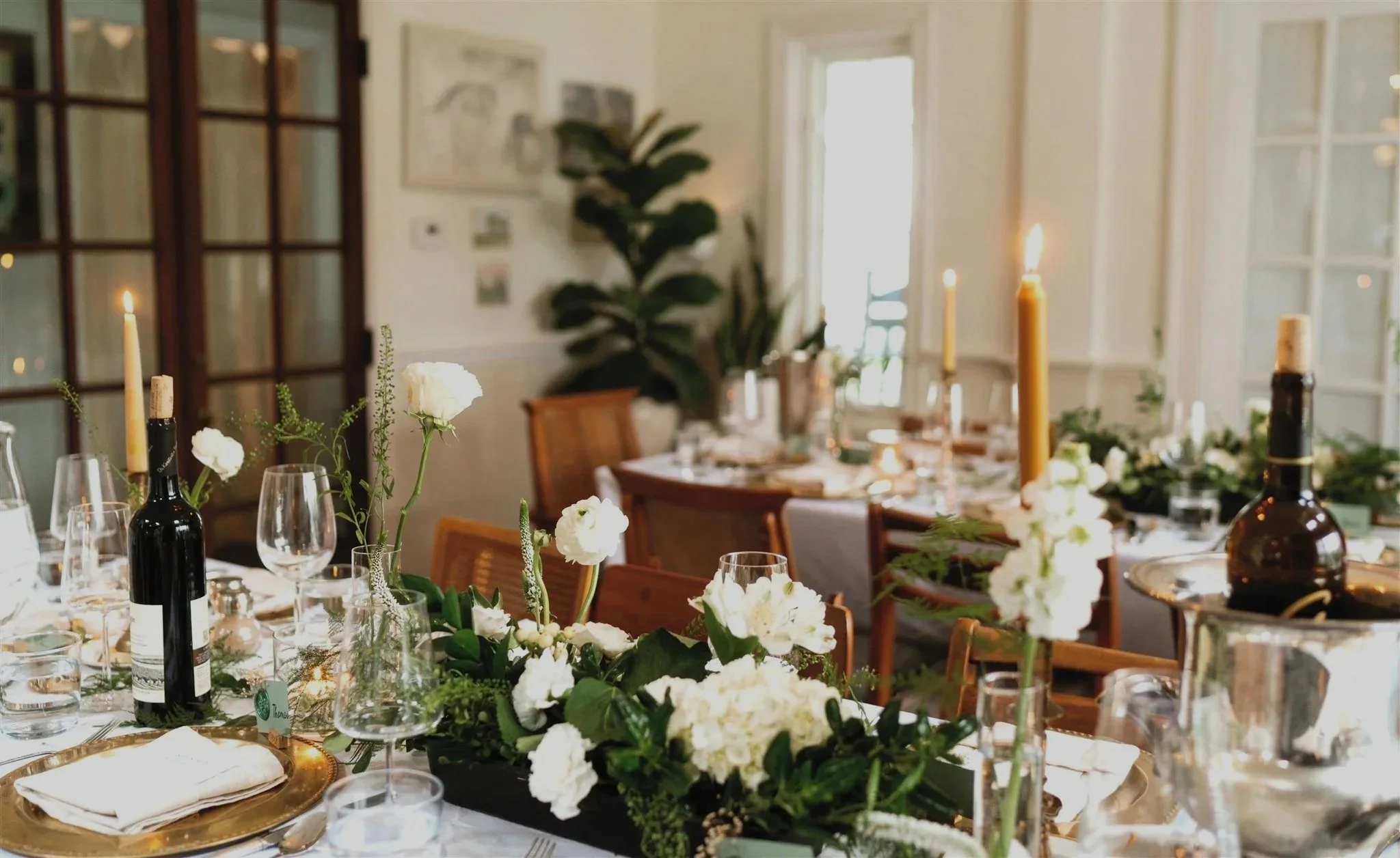 Elegant dining table setup with white tablecloth, gold chargers, green napkins, candles, and floral centerpieces featuring white flowers.