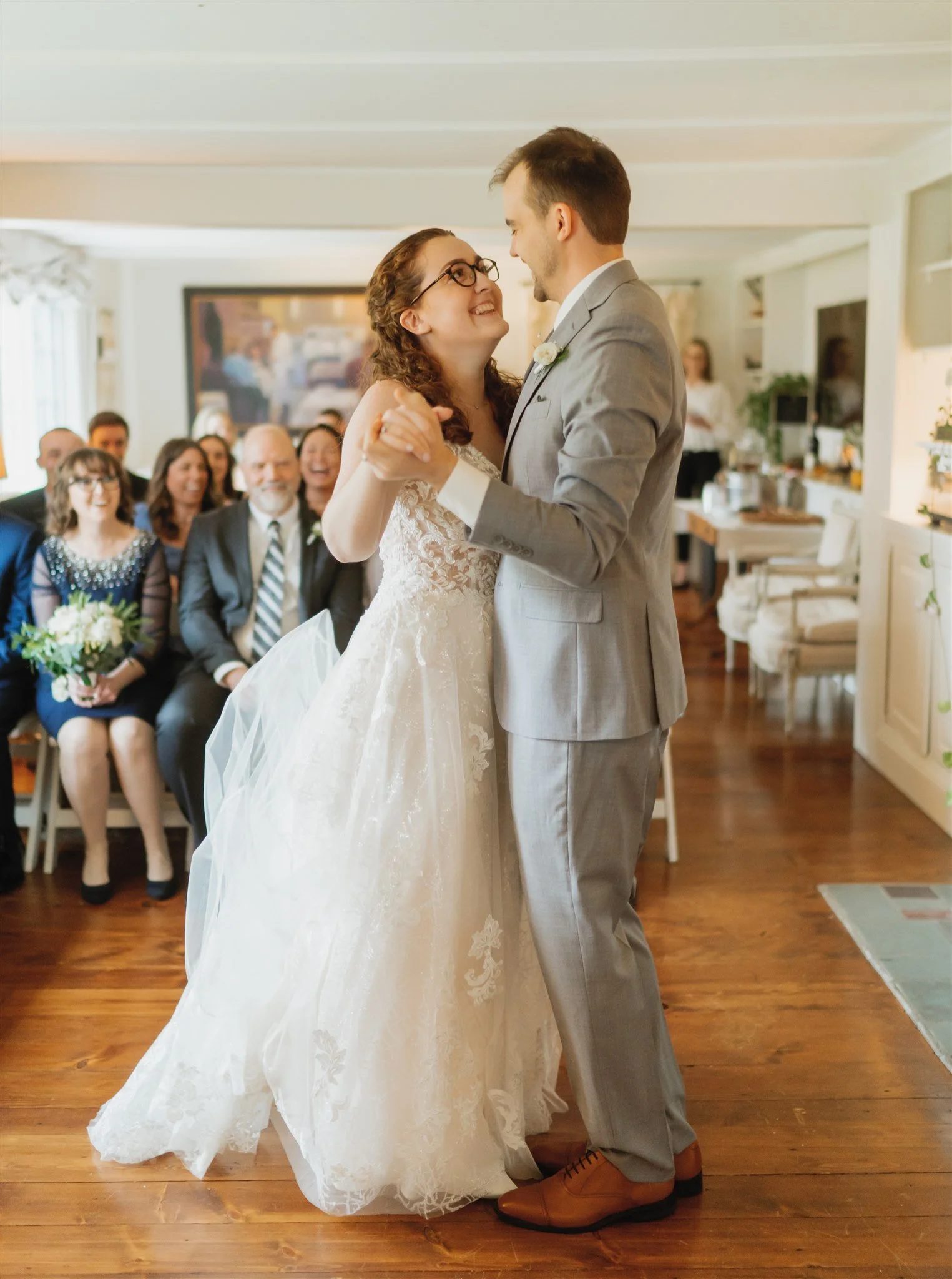 A bride and groom sharing their first dance at their wedding, surrounded by seated guests, in a warmly lit room with wooden flooring.