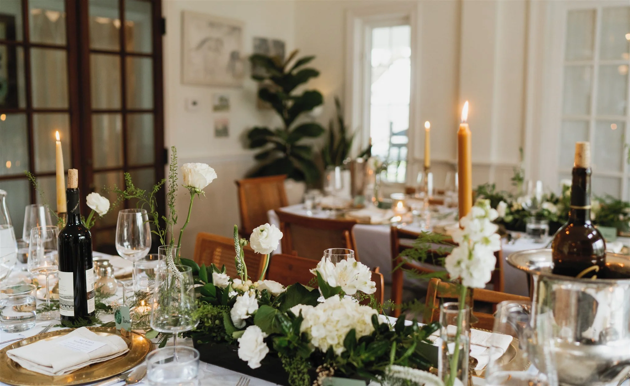 Elegant dining table setup with white tablecloth, gold chargers, green napkins, candles, and floral centerpieces featuring white flowers.