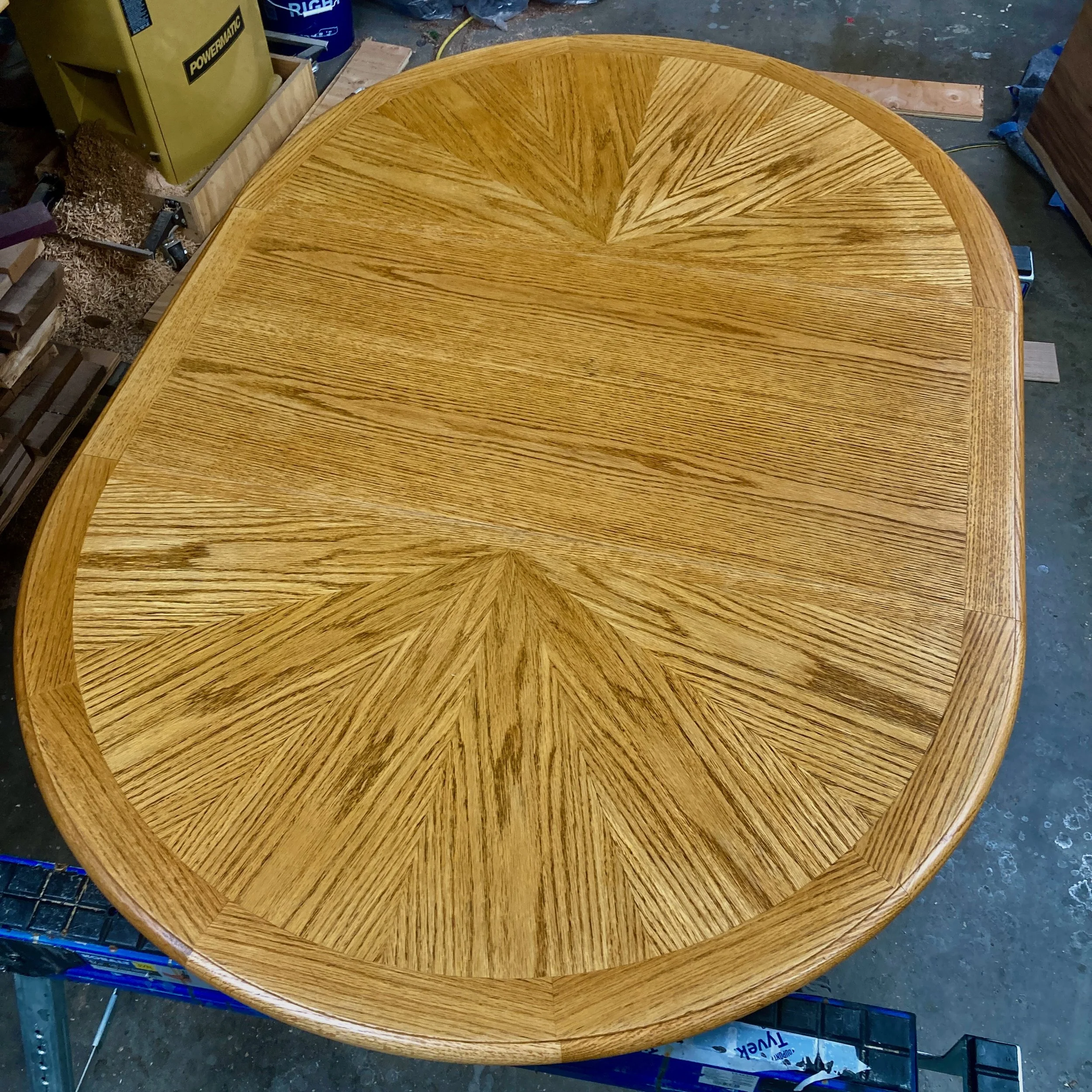 A round wooden table with a finished surface, featuring a parquet pattern with alternating wood grain directions in a woodworking workshop.