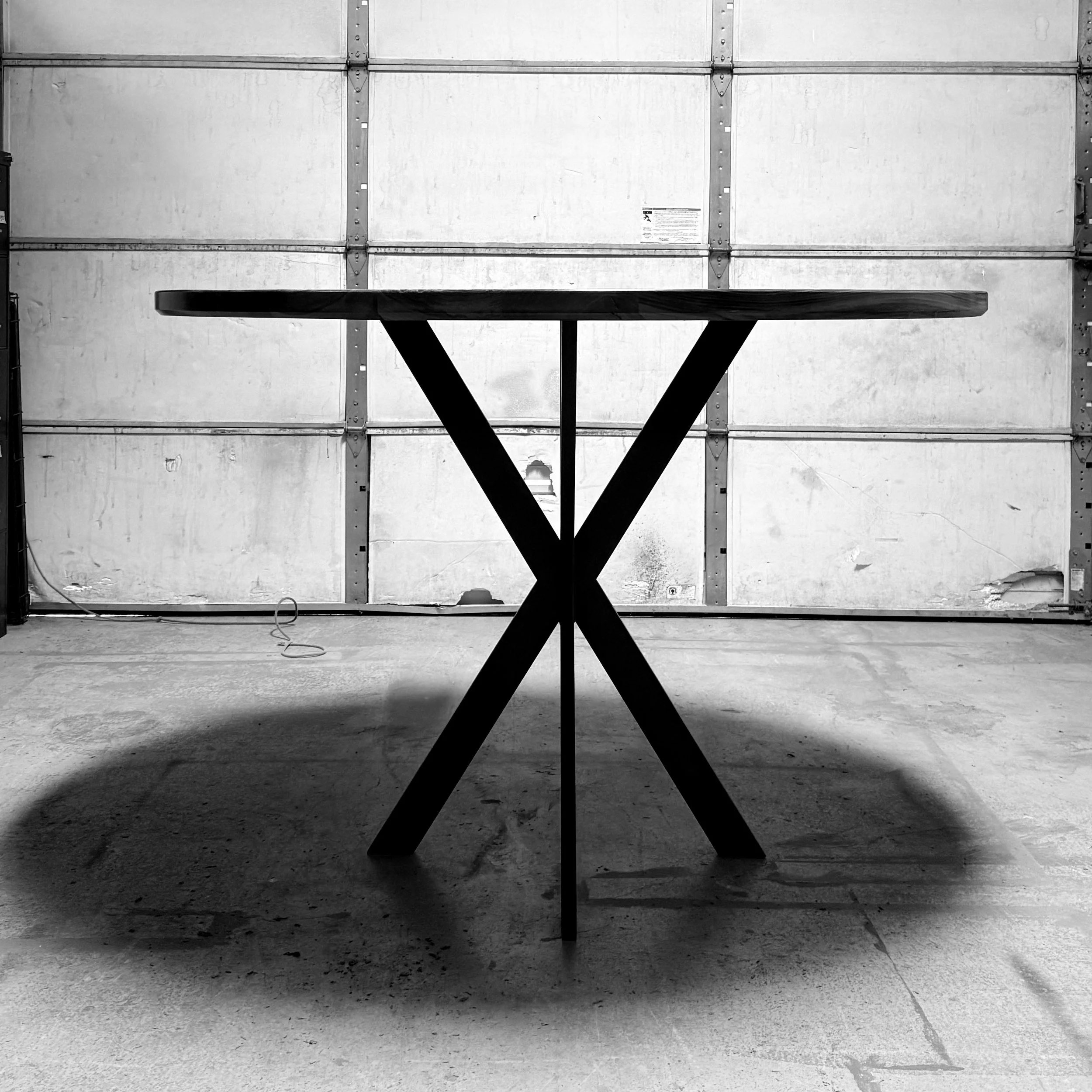 A round wooden table with crossed black legs is positioned in a garage with a closed door in the background.