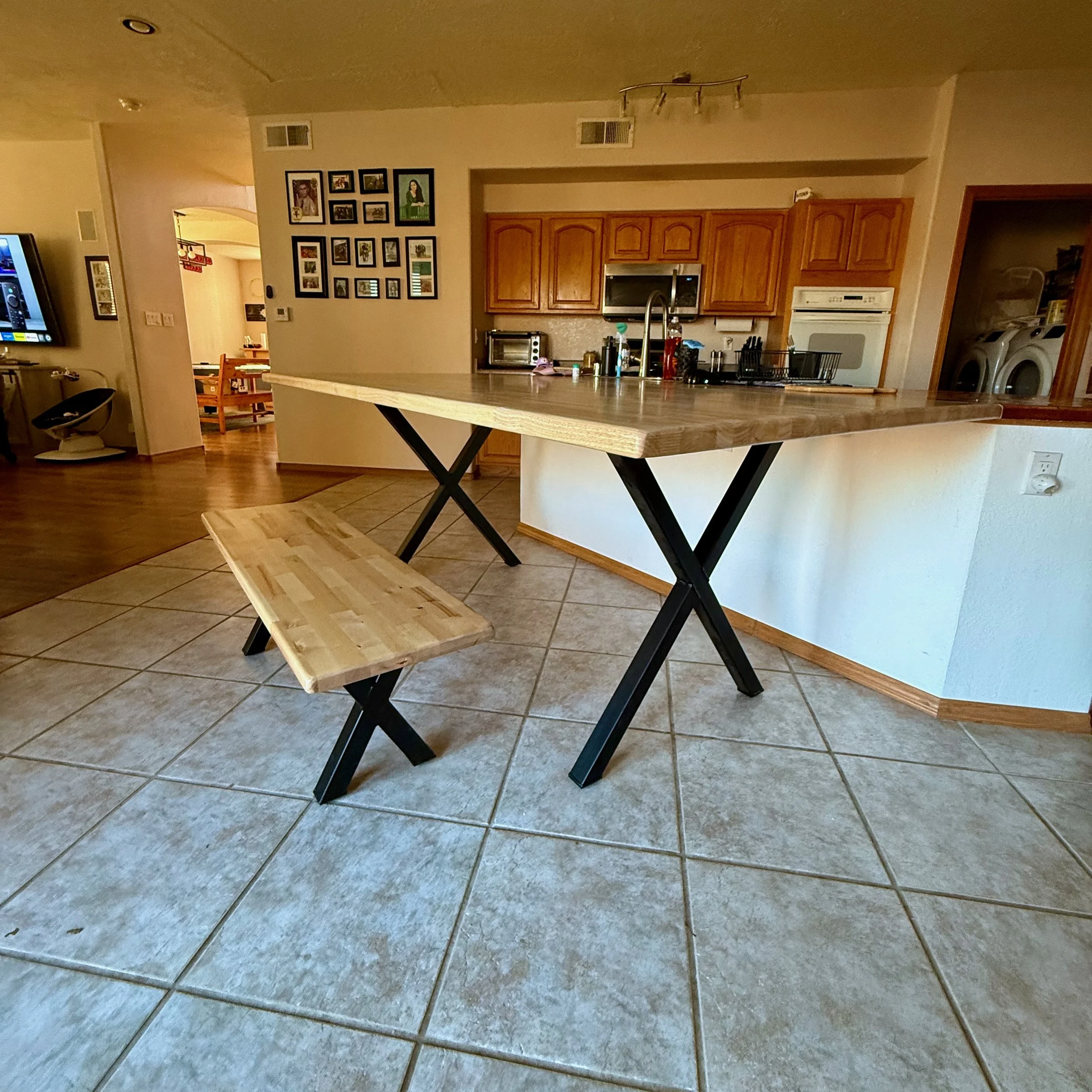 Kitchen with wooden cabinets, a countertop, hanging utensils, a washer and dryer in an adjacent room, and a breakfast bar with a wood top and black metal legs, along with a matching wooden bench.