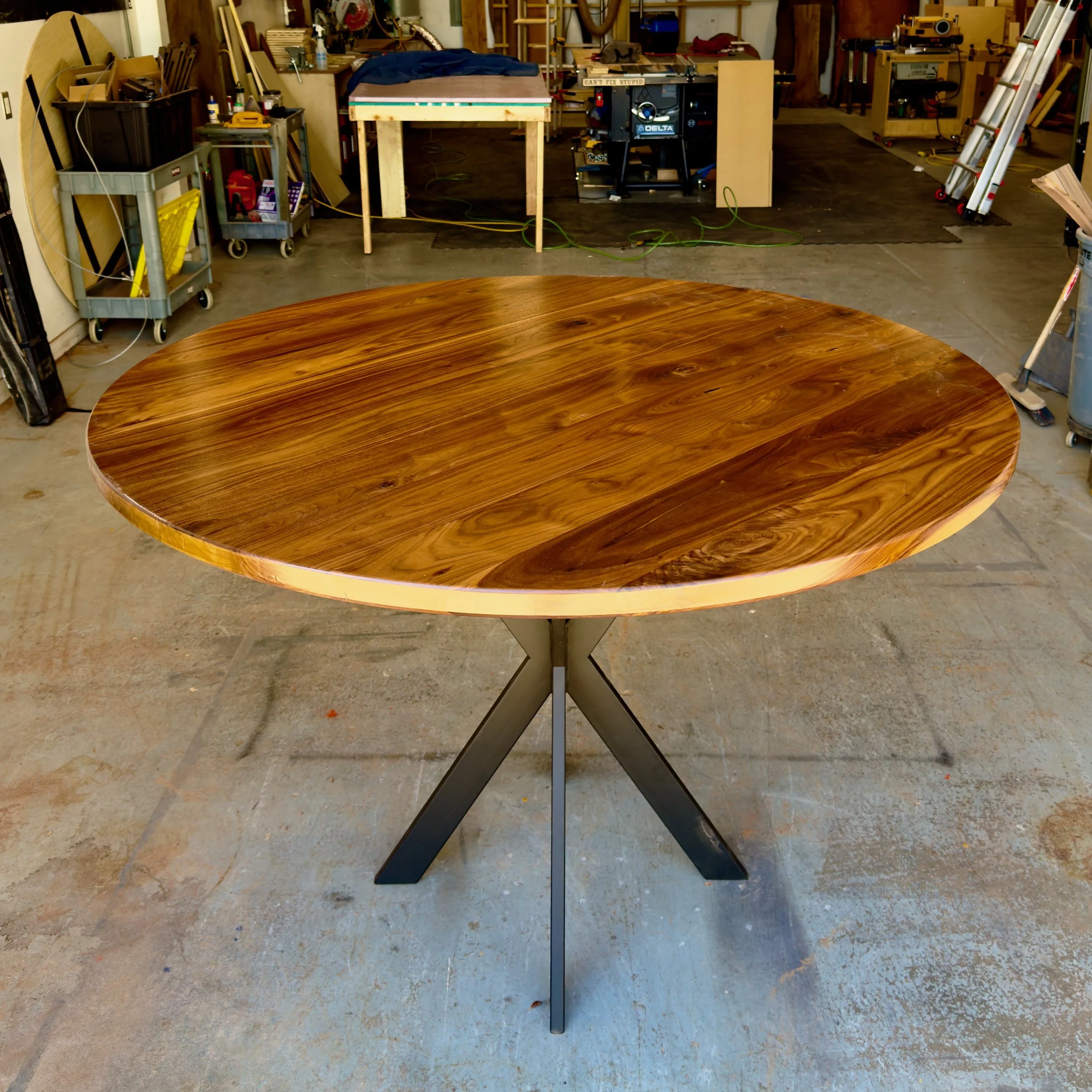 Round wooden table with a polished surface and dark metal legs, situated in a woodworking workshop with tools and equipment in the background.