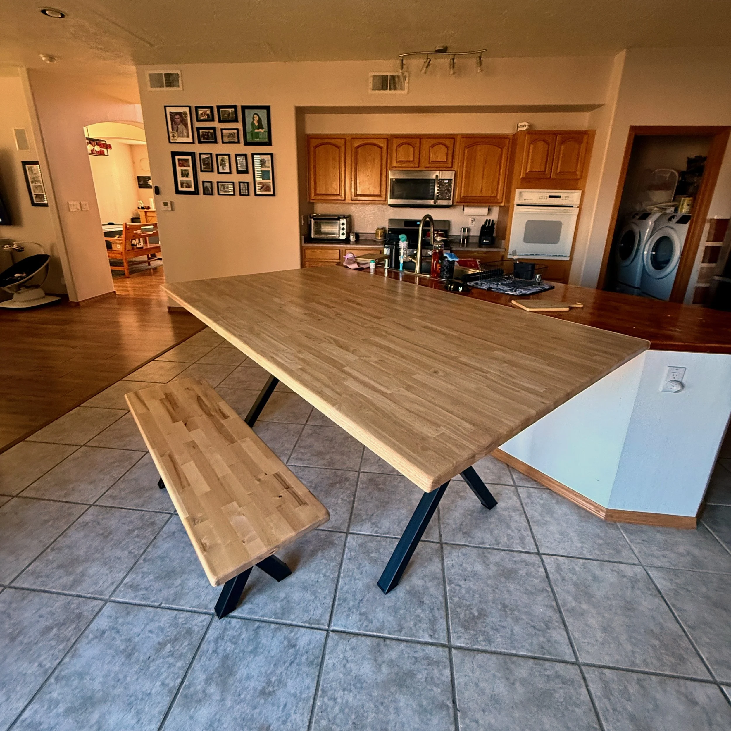 A wooden dining table with black metal legs and matching wooden bench in a kitchen area, with kitchen appliances and framed photos on the walls in the background.