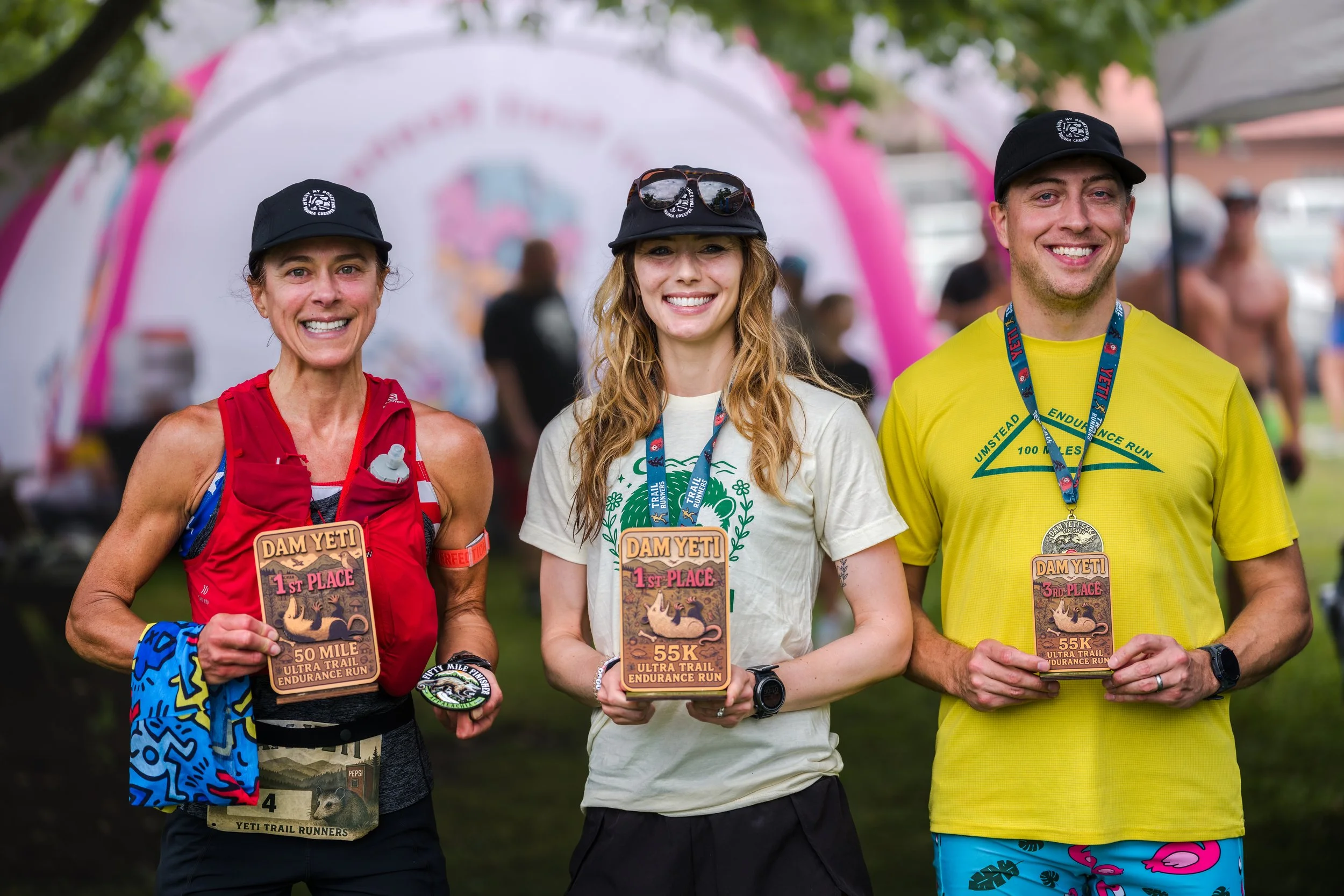 Three smiling runners holding race plaques after a trail run event, with a pink and white event tent in the background. All wear medals around their necks.