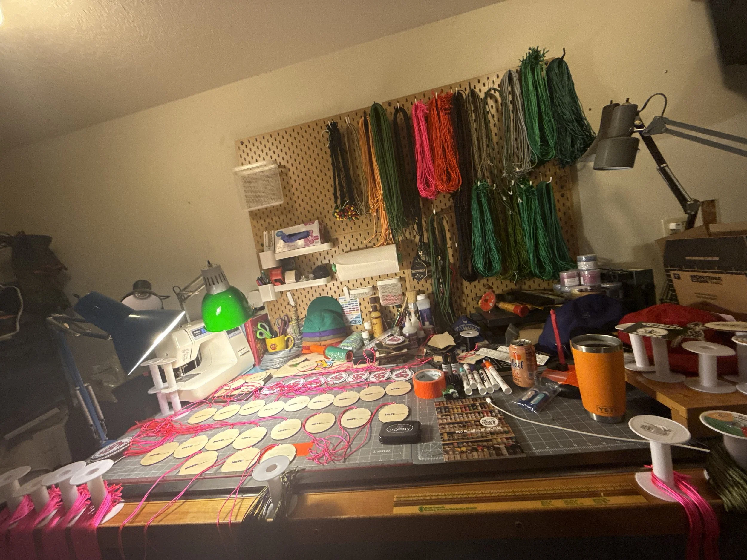 A cluttered work table with various crafting supplies, including multiple spools of pink thread, markers, scissors, and small containers. The background features a pegboard with colorful yarns or threads hanging and a desk lamp shining on the workspace.