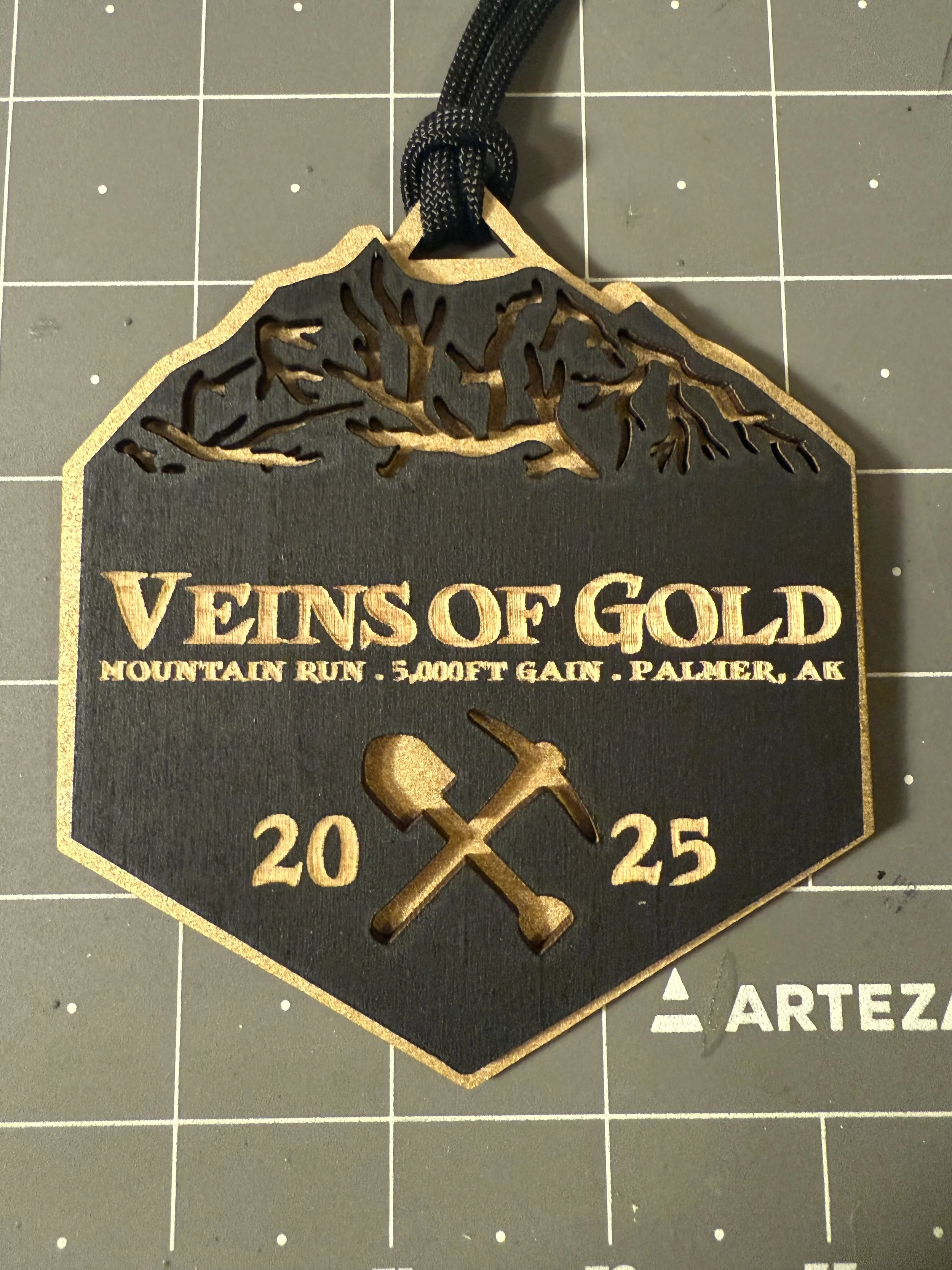 A black and gold commemorative plaque with a mountain and pickaxe design, reading "Veins of Gold, Mountain Run, 5000 Feet Gain, Palmer, AK, 2025, 20, 25."