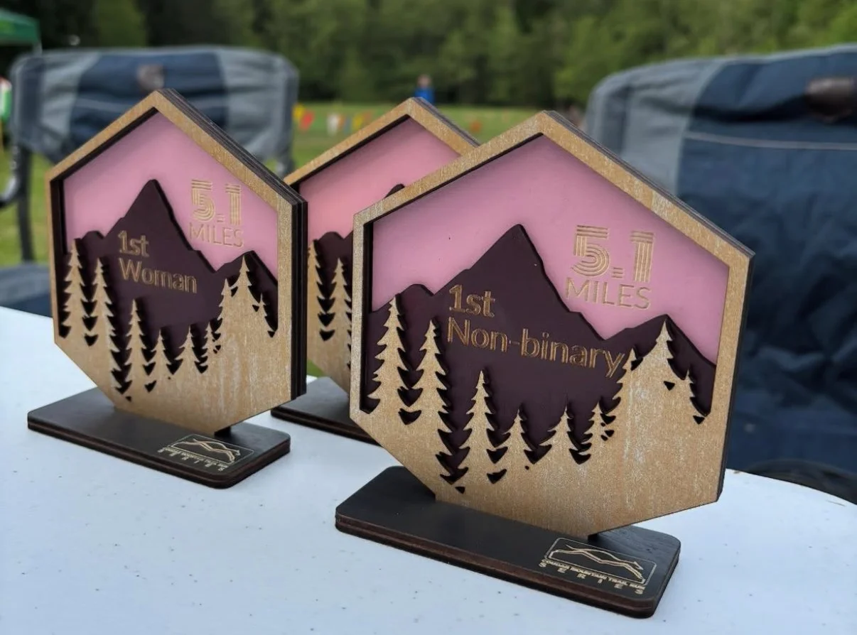 Wooden awards with mountain and pine tree design, engraved with '51 miles,' '1st Woman,' and '1st Non-binary,' set on a white surface outdoors.