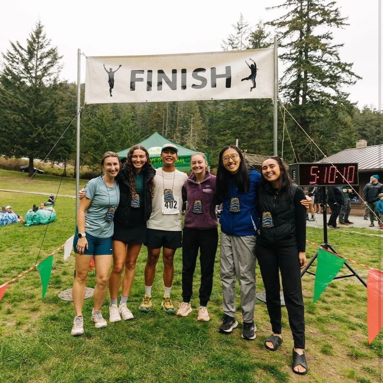 Group of six women celebrating crossing the finish line of a race outdoors, with a large finish banner and a digital clock showing 51 minutes. The women are smiling and wearing athletic clothing and medals, standing on a grassy area surrounded by tre