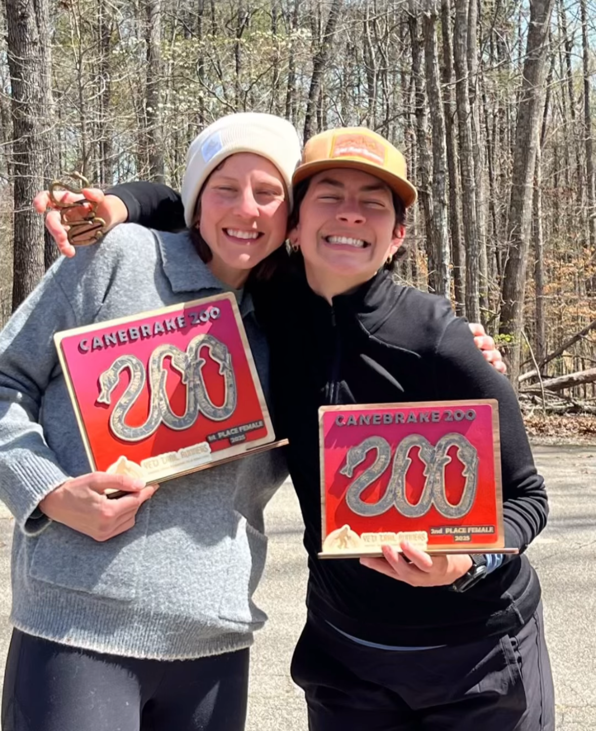 Two smiling women holding awards shaped like snakes, celebrating their victory at CaneBarke Zoo, with a wooded area in the background.