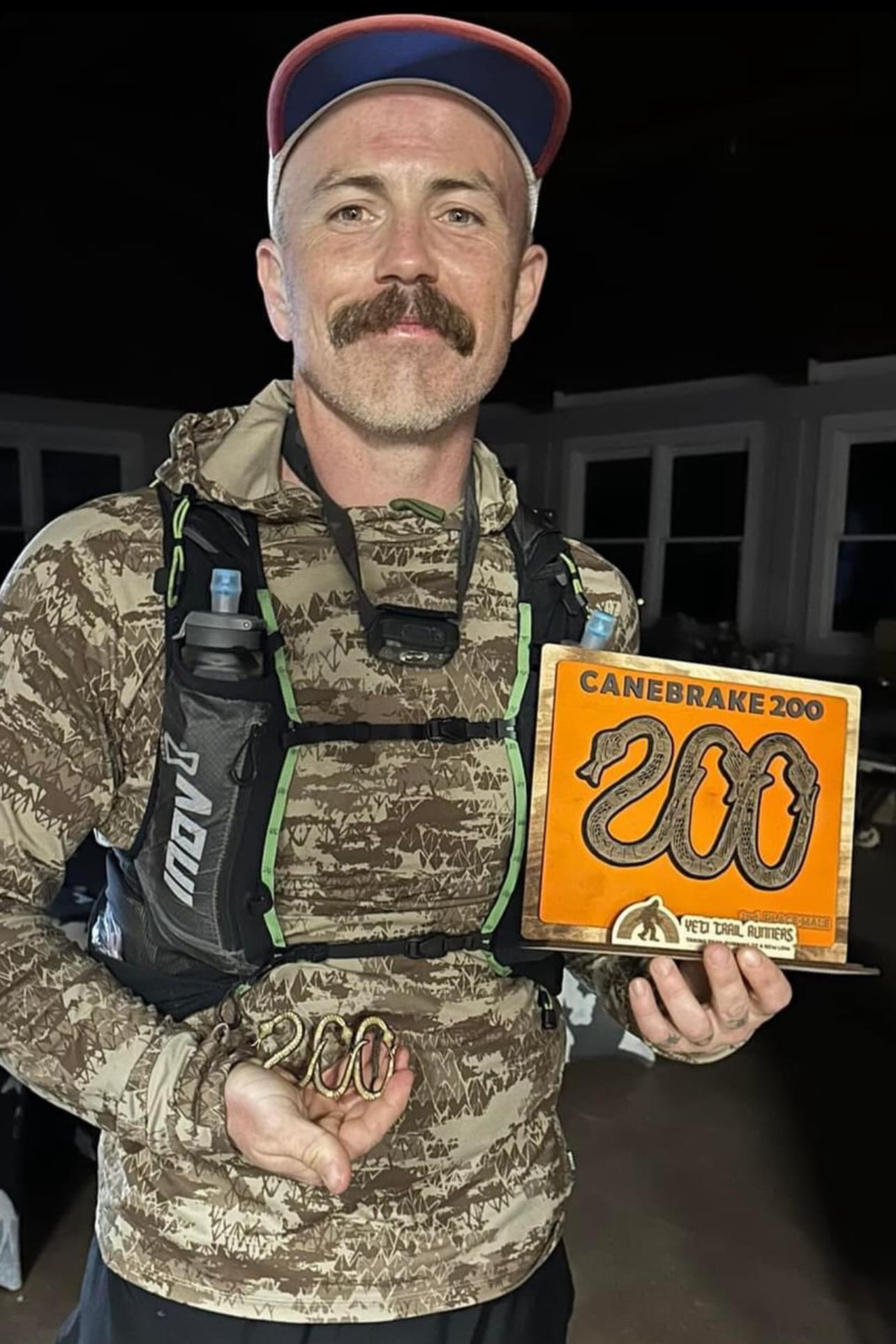 A man with a mustache and beard smiling, wearing a camouflage-patterned jacket and a red and blue cap, holding a numbered sign and a metal snake-shaped object.