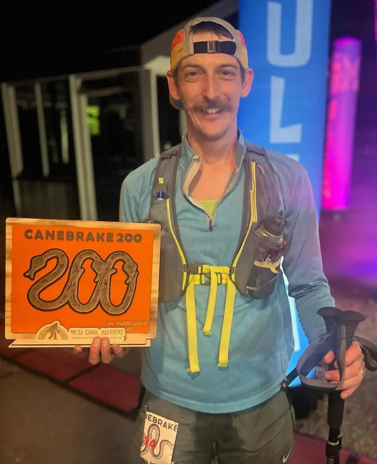 Man smiling, wearing a cap backwards, holding a finish line sign with the number 200, wearing a running vest and holding a running pole, after a trail race.