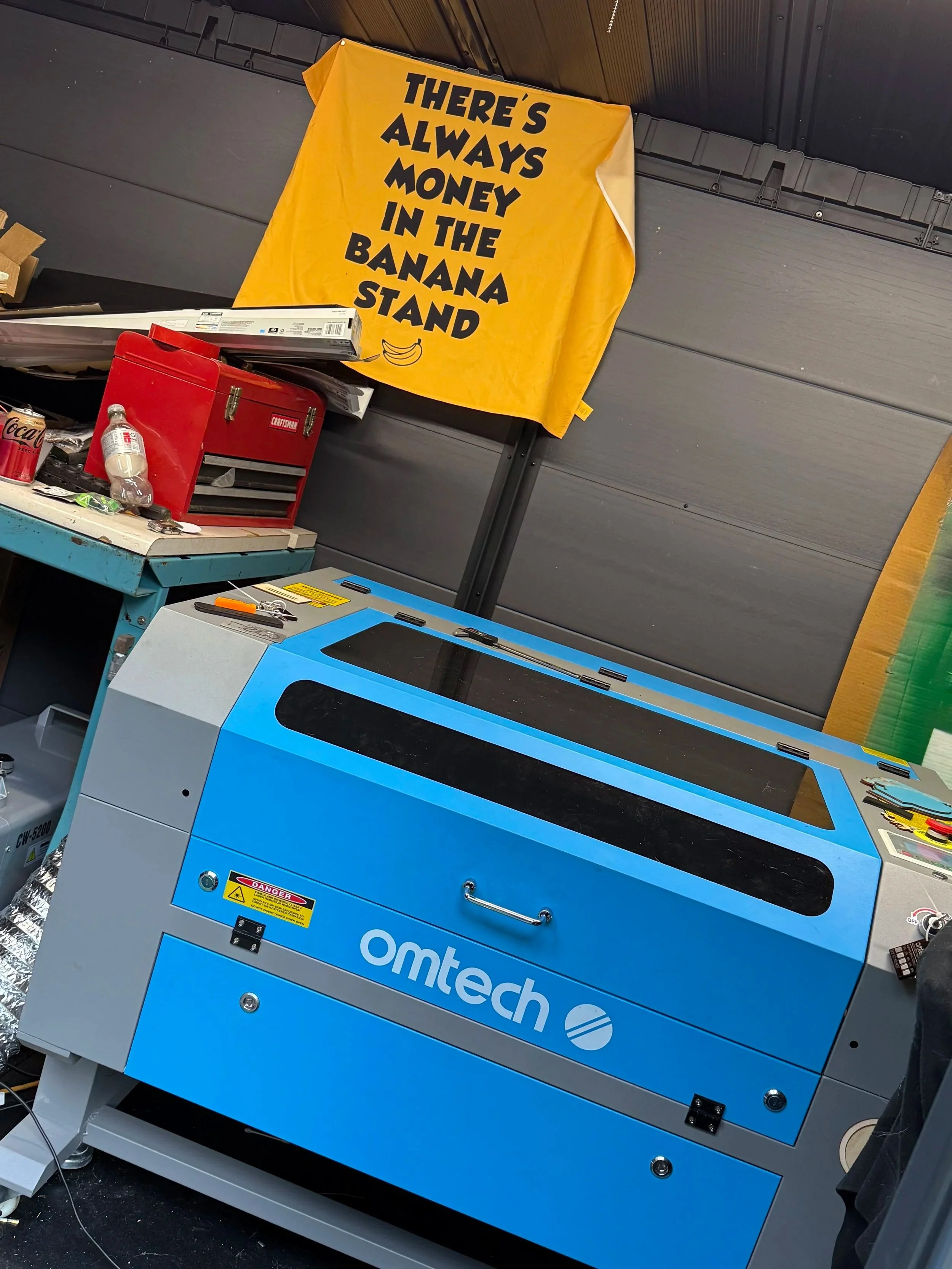 A blue OMTech laser engraver in a workshop, with a yellow fabric banner hanging above that reads, 'There's always money in the banana stand,' with a small banana graphic below the text.