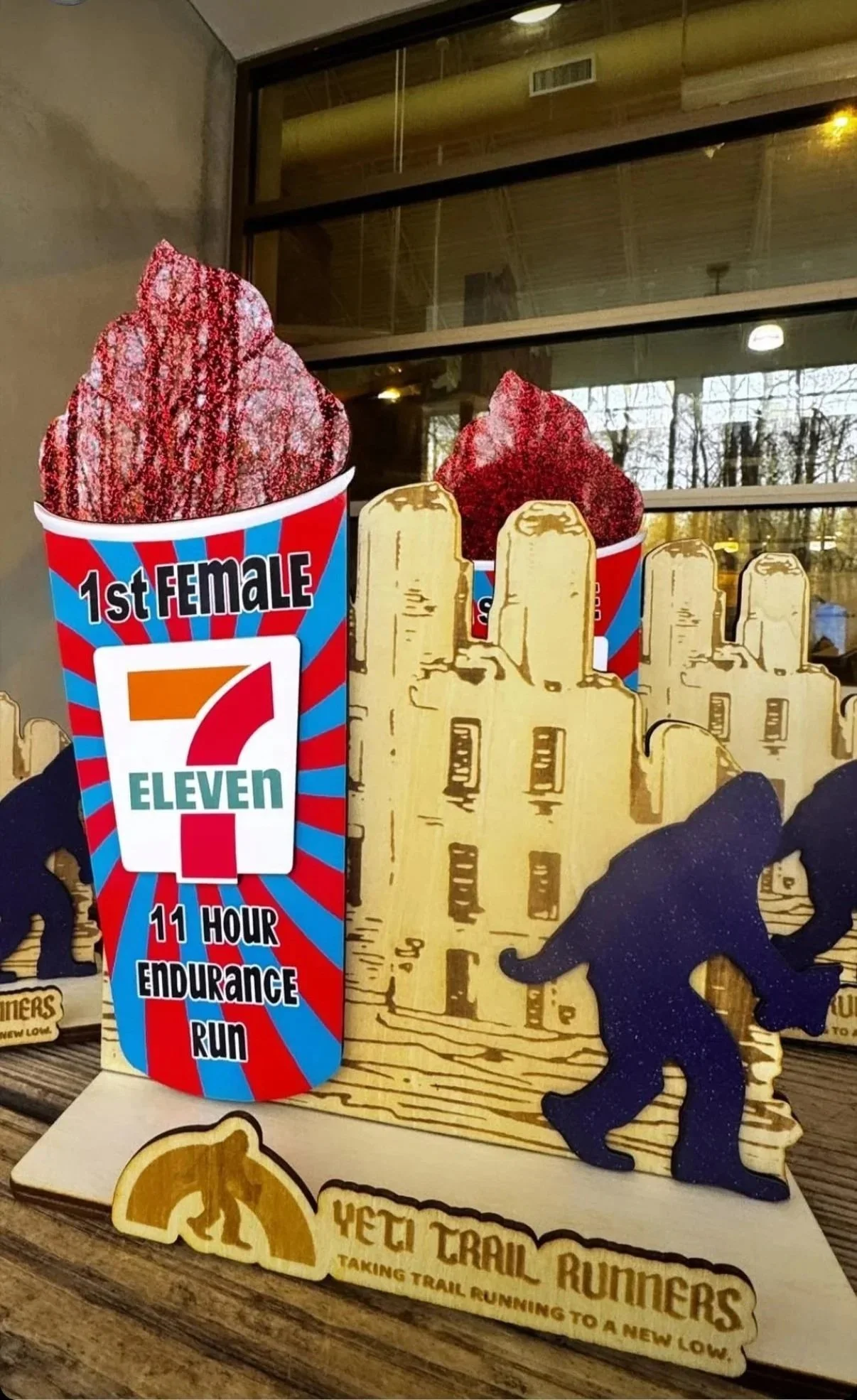 Decorative display featuring wooden cutouts of a city skyline, a runner silhouette, and a sign that says "YETI TRAIL RUNNERS TAKING TRAIL RUNNING TO A NEW LOW," with two cups of frozen red velvet treat labeled "1st FEMALE 7 ELEVEN 11 HOUR ENDURANCE R