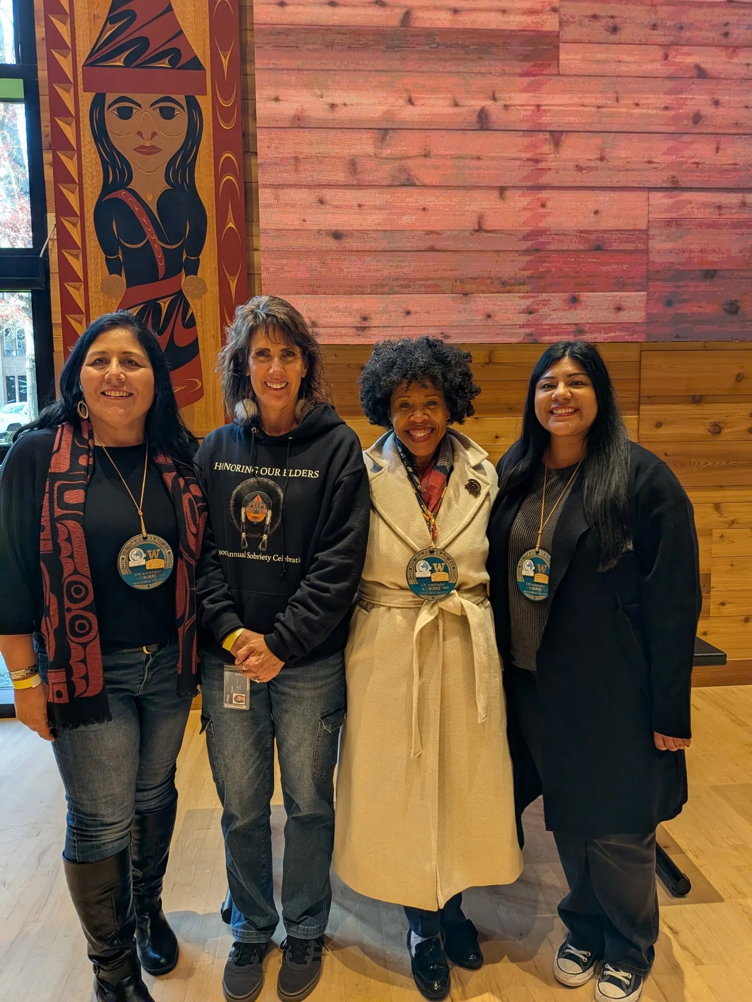 Four women standing together in a room with wooden walls and artwork, smiling at the camera, wearing conference badges.