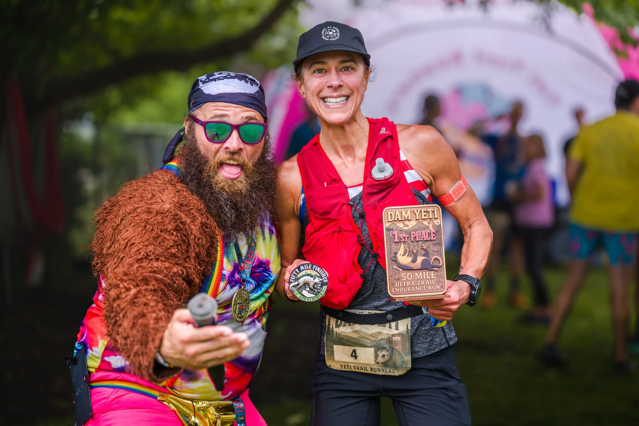 Two runners celebrating after completing a 50-mile ultra trail race. The man on the left has a bushy beard, wearing sunglasses, a headband, and a colorful shirt, holding a microphone and wearing a medal. The woman on the right is smiling, wearing a black cap, a red running vest, and holding a plaque that says 'Dam Yeti, 1st Place, 50 Mile Ultra Trail Endurance Run.' She has a race bib with the number 4 and appears to be smiling proudly.