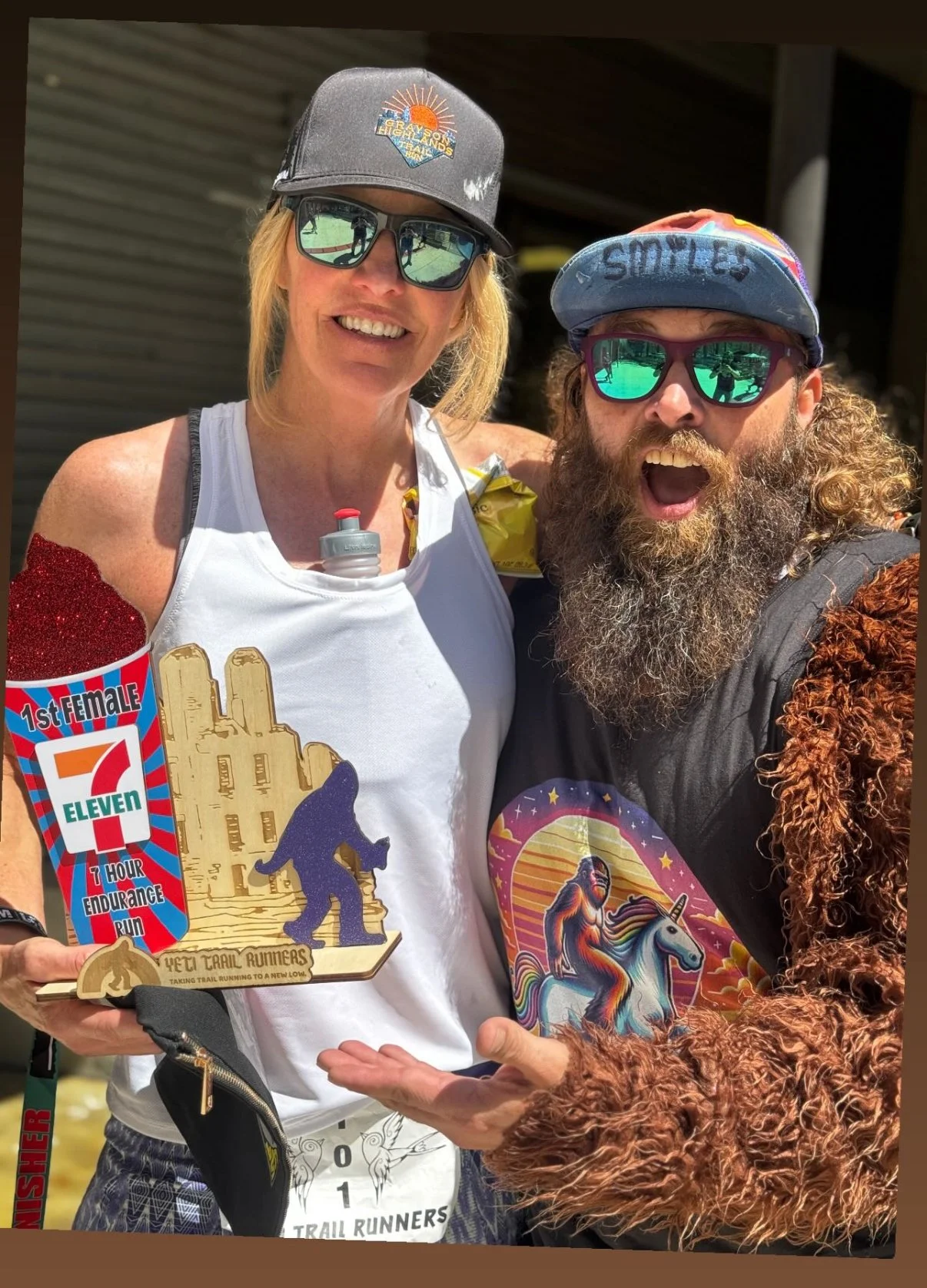 Two people celebrating after a trail running event, holding a wooden trophy shaped like a gorilla and a vertical sign with the 7-Eleven logo, both wearing sunglasses and casual athletic clothing, outdoors with a building in the background.