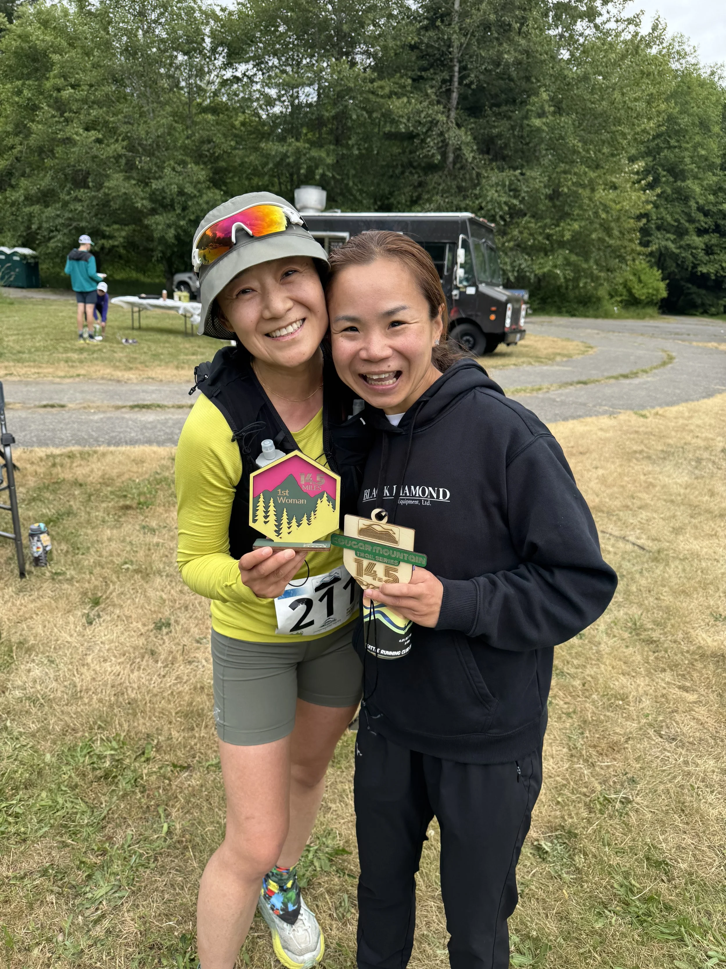 Two women smiling and holding race medals outdoors, with a food truck and trees in the background.