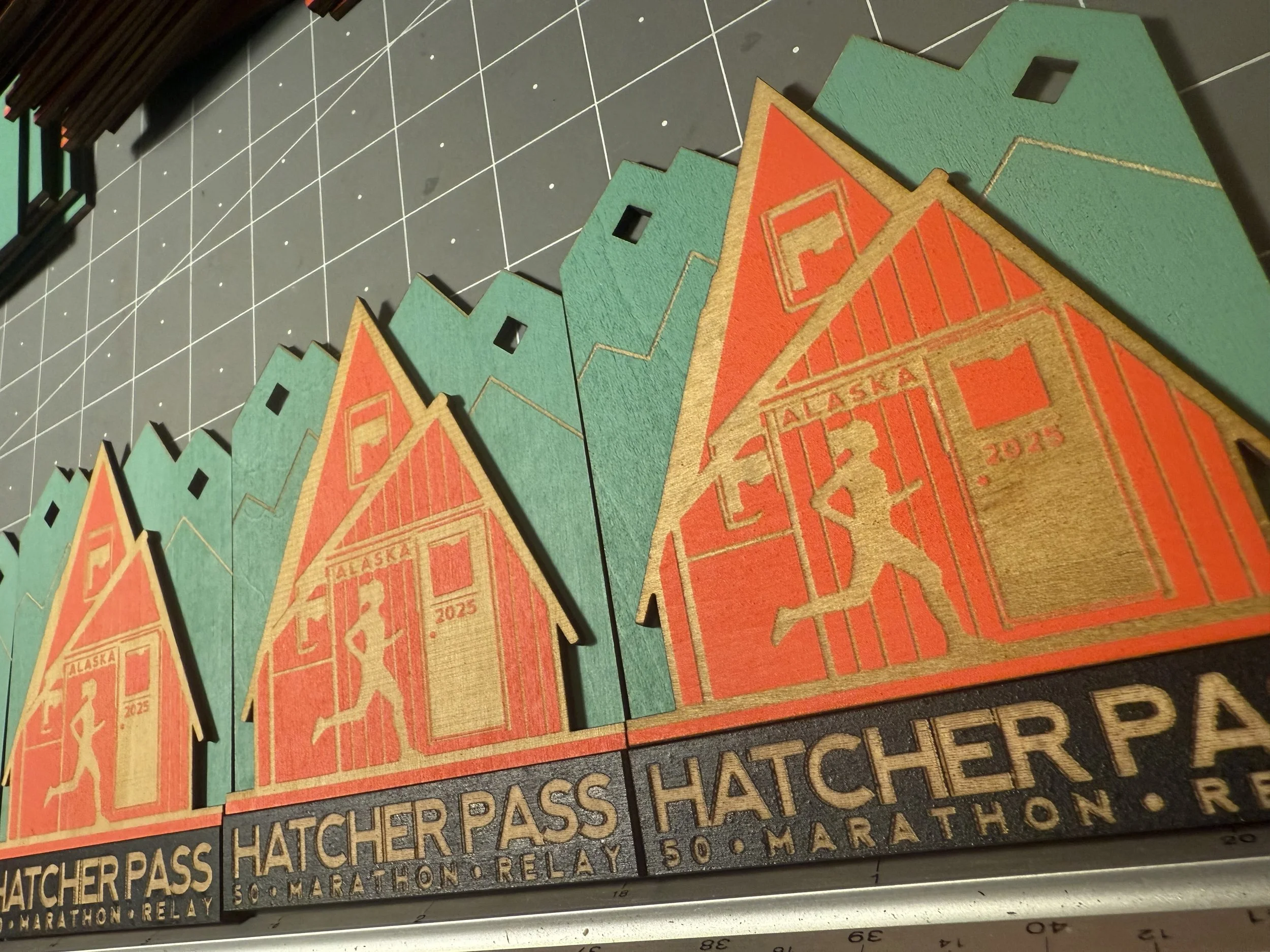 Decorative wooden plaques for the Hatcher Pass 50th Marathon Relay featuring mountain peaks, a hiker silhouette, and the text 'Alaska 2025'.