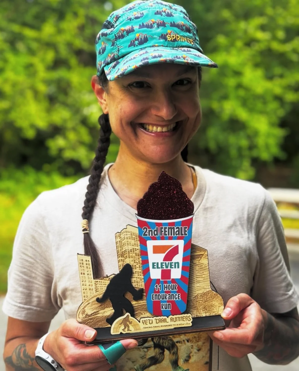 A woman smiling, holding a trophy with a cup filled with a dark, glittery substance, featuring a label that says '2nd Female 7 Eleven 11 Hour Endurance Run,' with a background of green trees.