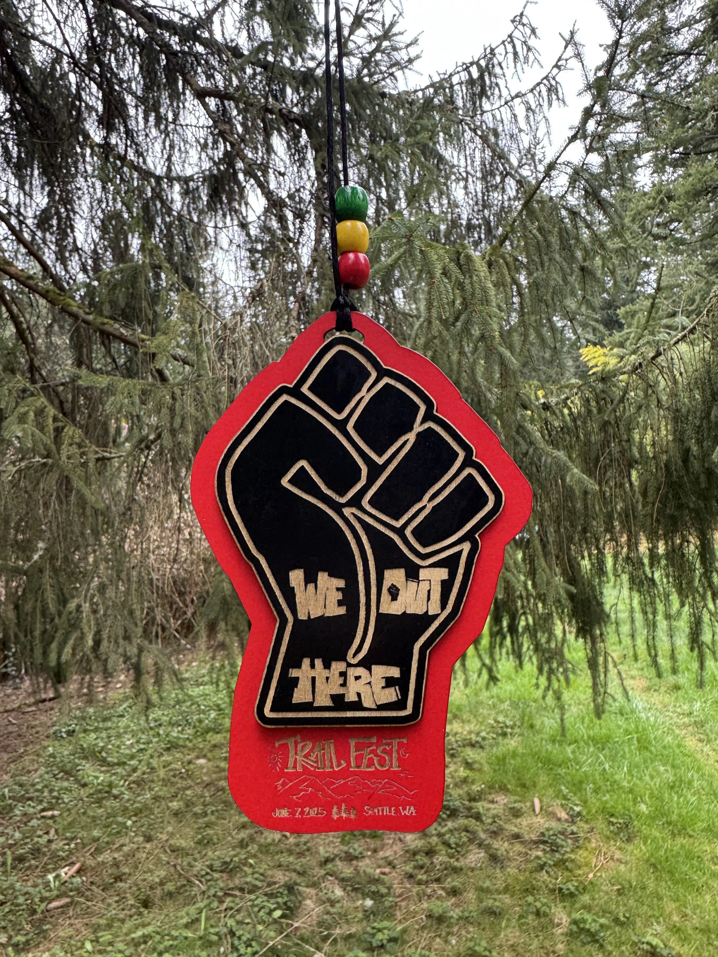 A red foam sign with a black fist and the text "We Out There" hangs from a tree, with beads on the string above it. The background shows green grass and tree branches.