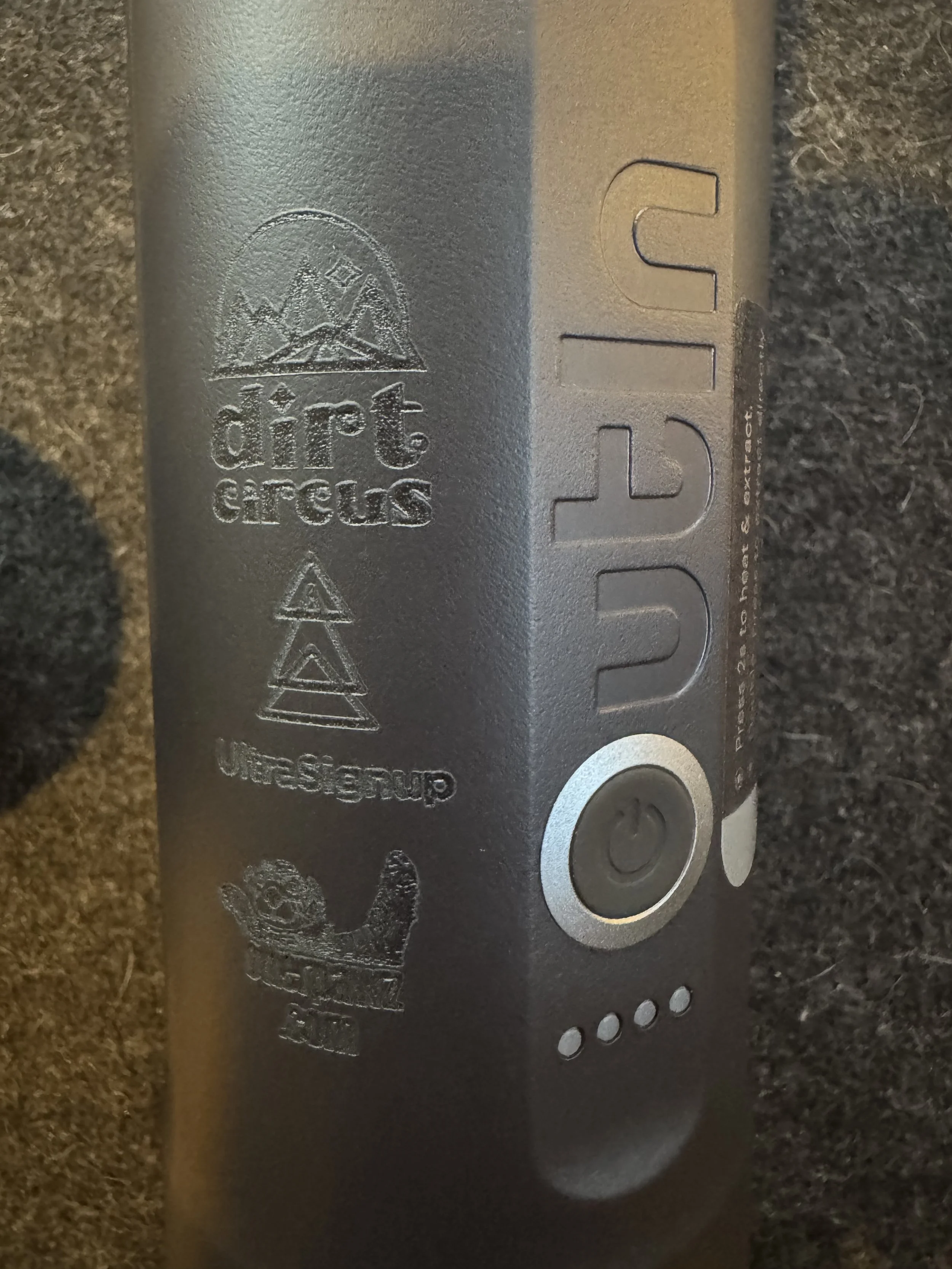 Close-up of a gray UlteStrom electric device with a power button and four indicator lights, placed on a dark surface. The device has an embossed logo and text.