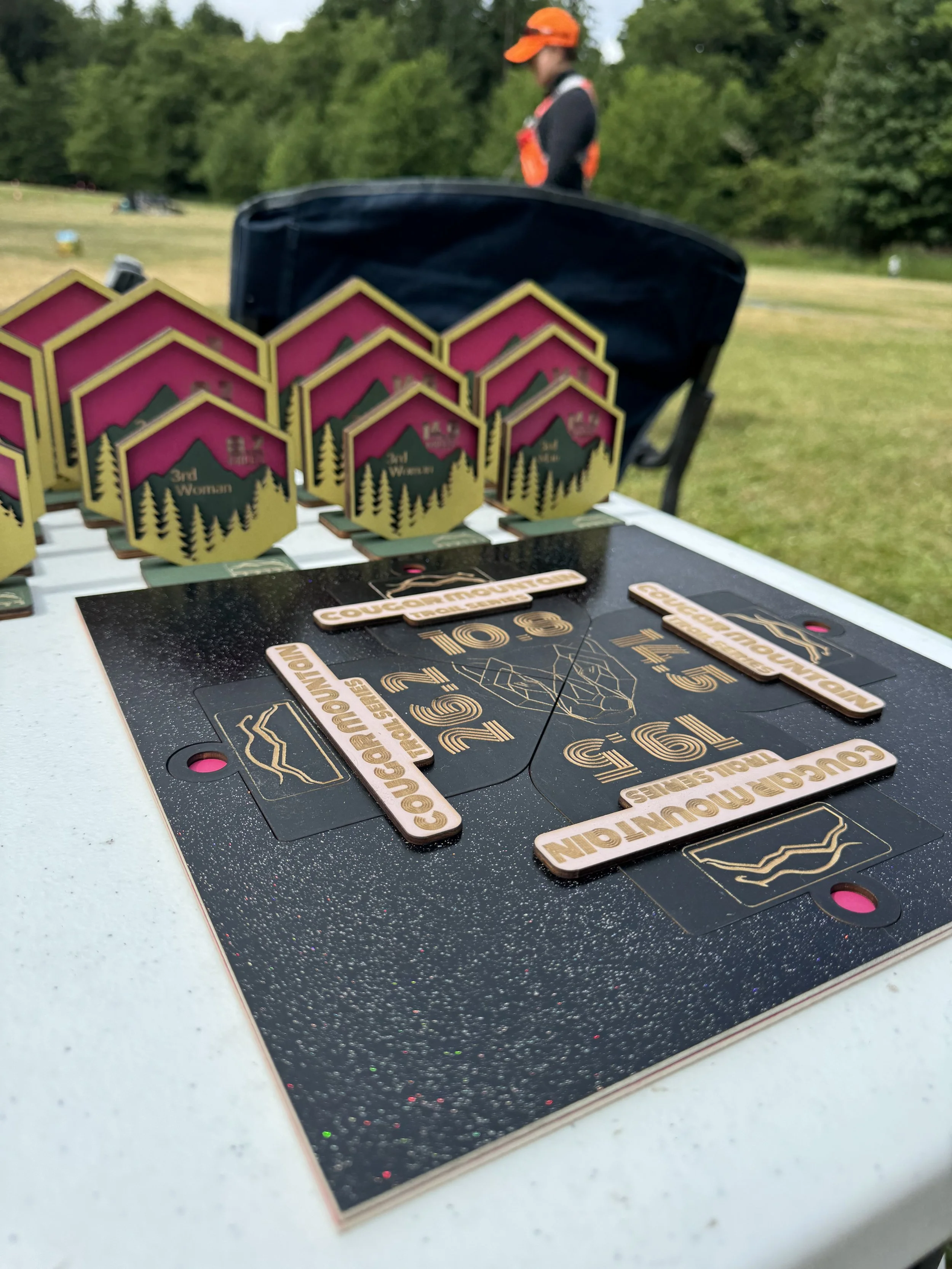 Golf course with a table displaying tournament awards, including medals and wooden plaques, in front of a person wearing an orange cap and black shirt.