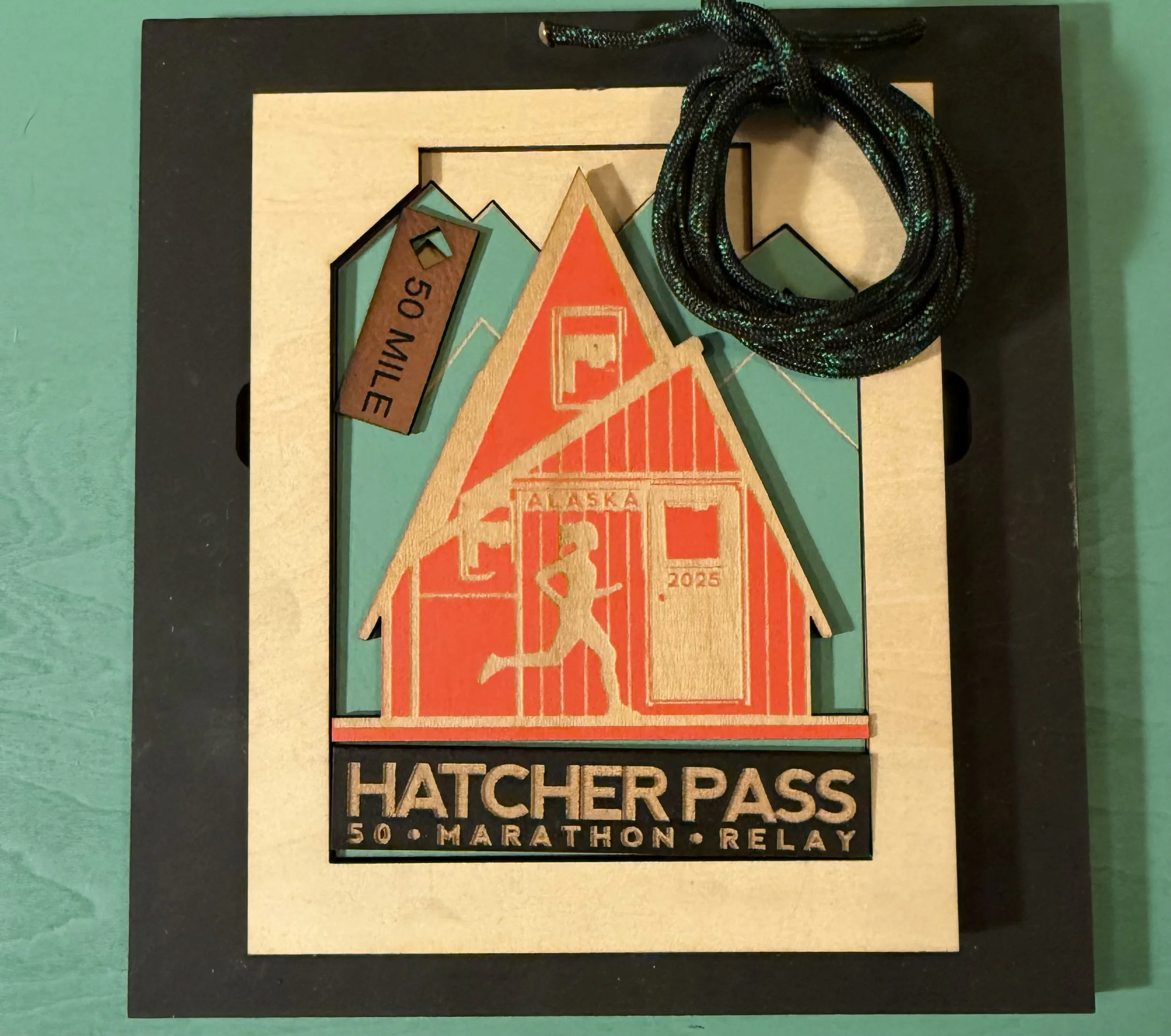 A framed memento from the Hatcher Pass 50-Mile Marathon Relay, featuring a wooden cutout of a mountain cabin with Alaska written on it, a silhouette of a runner, a watch, and a coiled black and green strap.