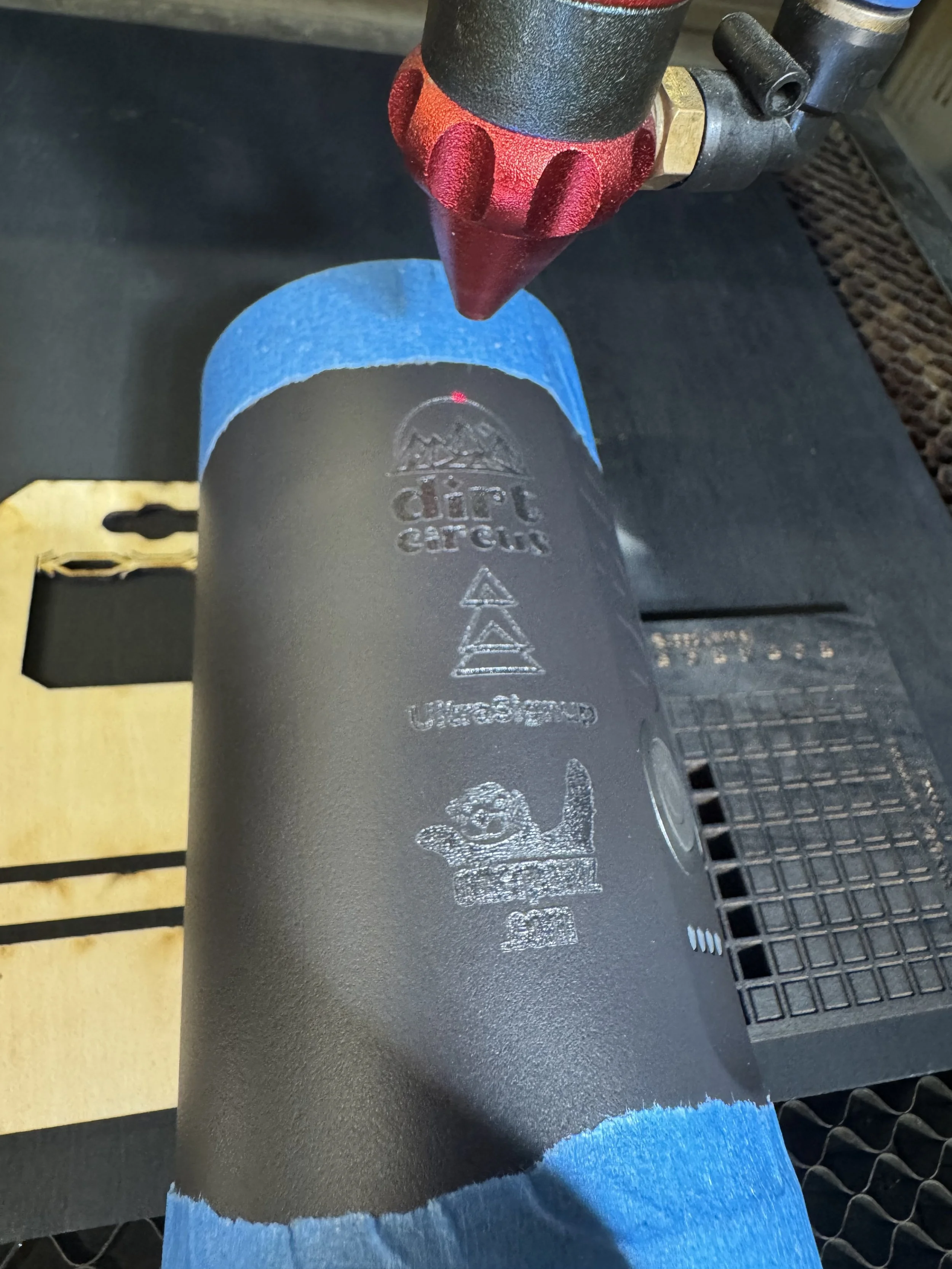 Laser engraving process on a black cylindrical surface with blue painter's tape around its edges.