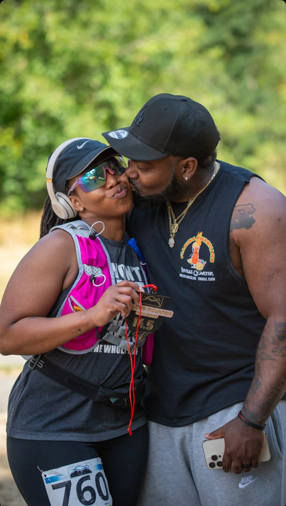 A woman and man share a kiss outdoors. The woman is wearing a black cap, sunglasses, a pink race bib, and athletic clothing, and is holding a medal. The man is wearing a black cap, sleeveless shirt, and is holding a phone.