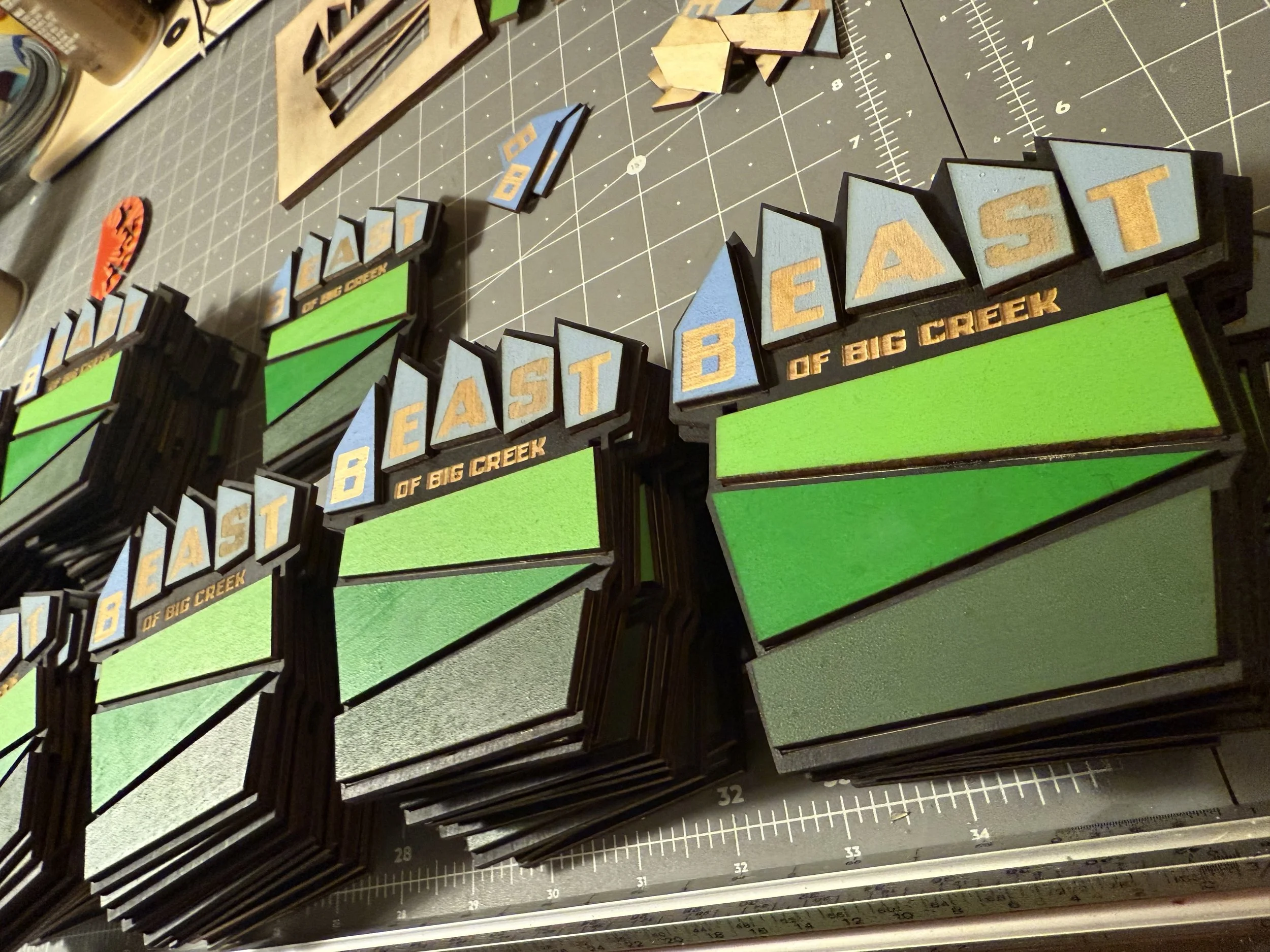 Multiple custom-made signs with the words "East Bast of Big Creek" arranged on a work table. The signs have blue, green, and black colors with layered lettering.