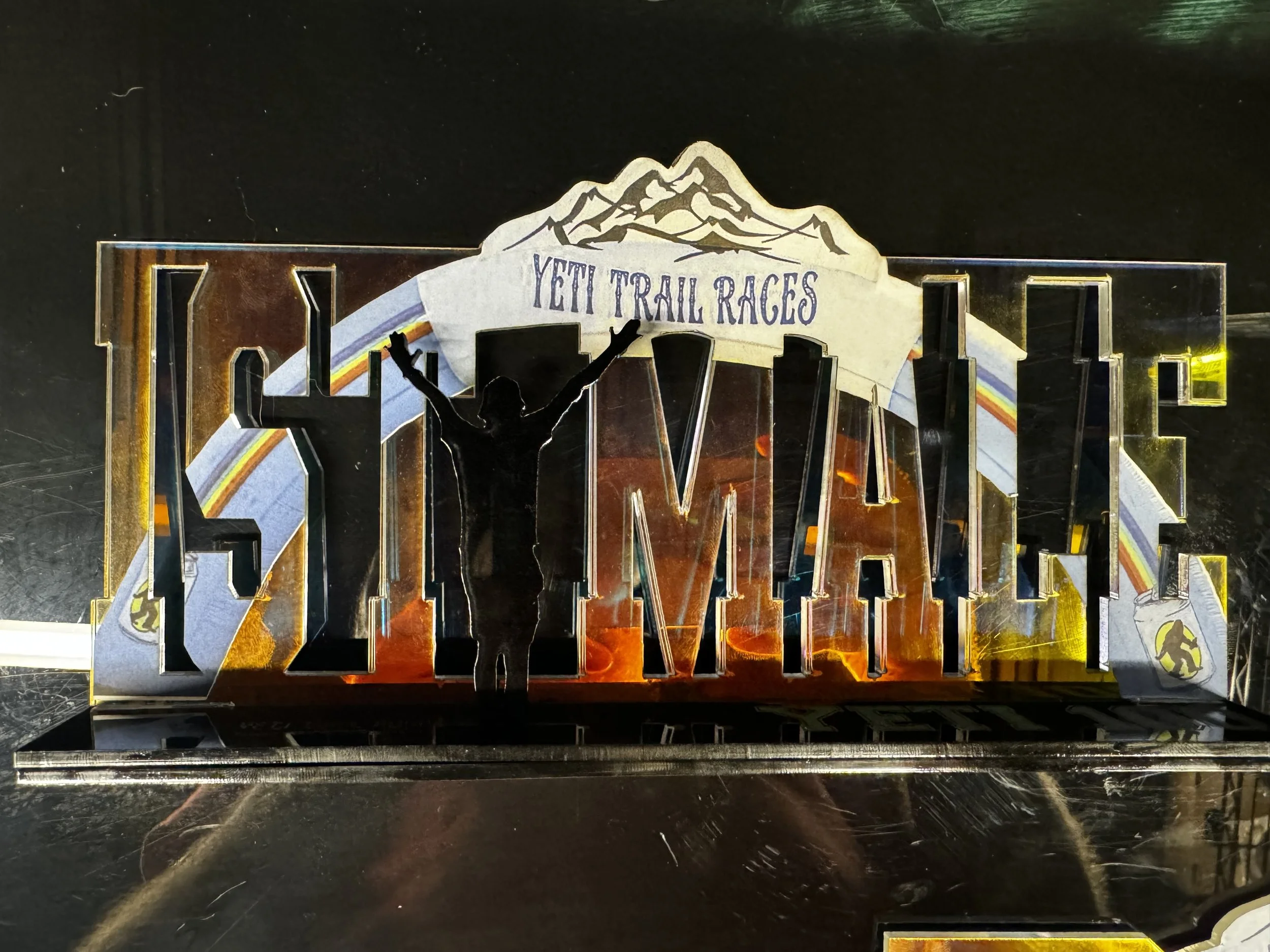 Metal sign with the words "HOME" and an outline of a person with arms raised, with a mountain and "YETI TRAIL RACES" banner in the background.