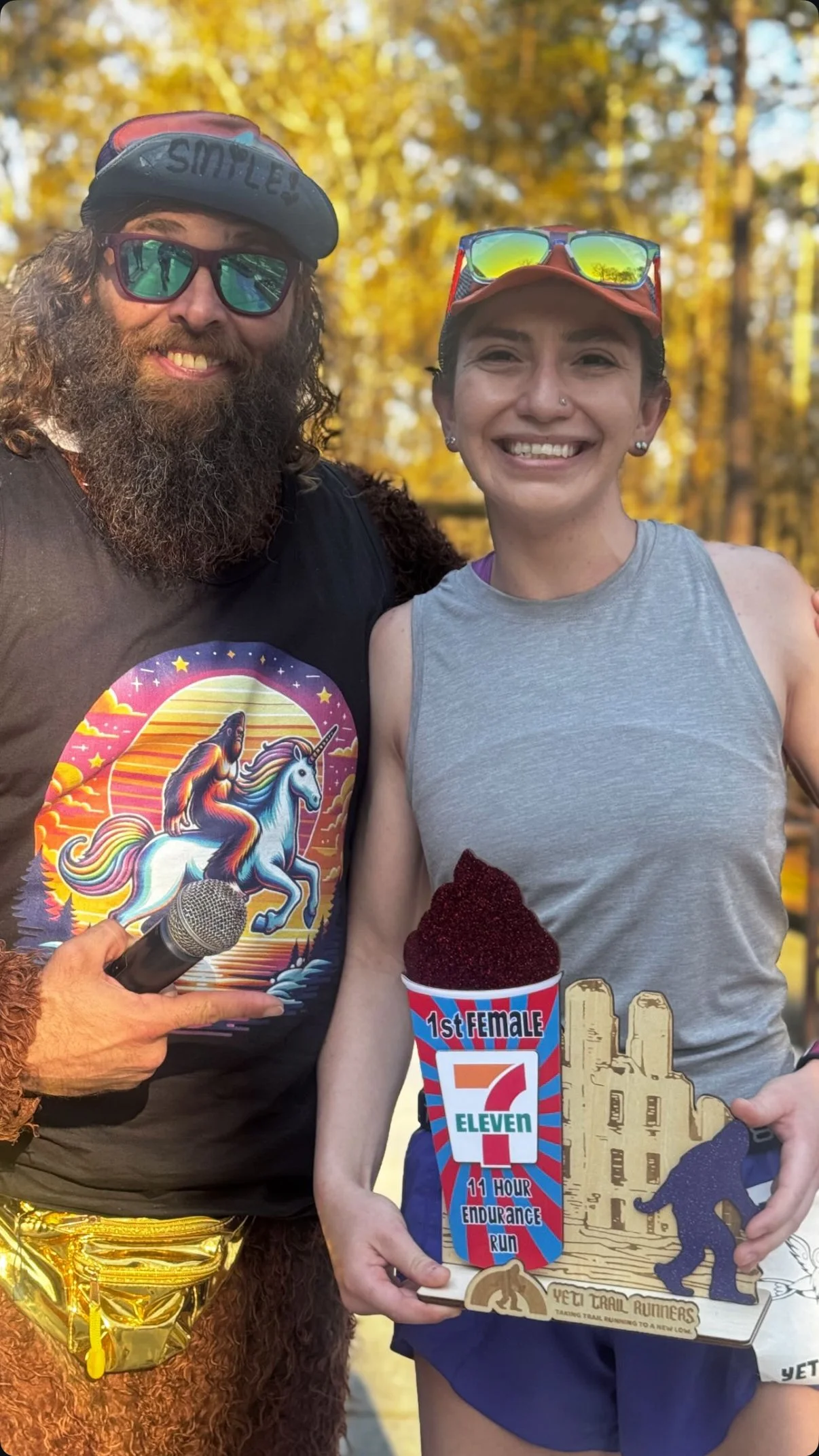 A bearded man in a black T-shirt with a colorful unicorn riding a rainbow and a woman in a gray tank top holding a wooden trail running award, with a large cup of red ice cream in front of her, standing outdoors with autumn trees in the background.
