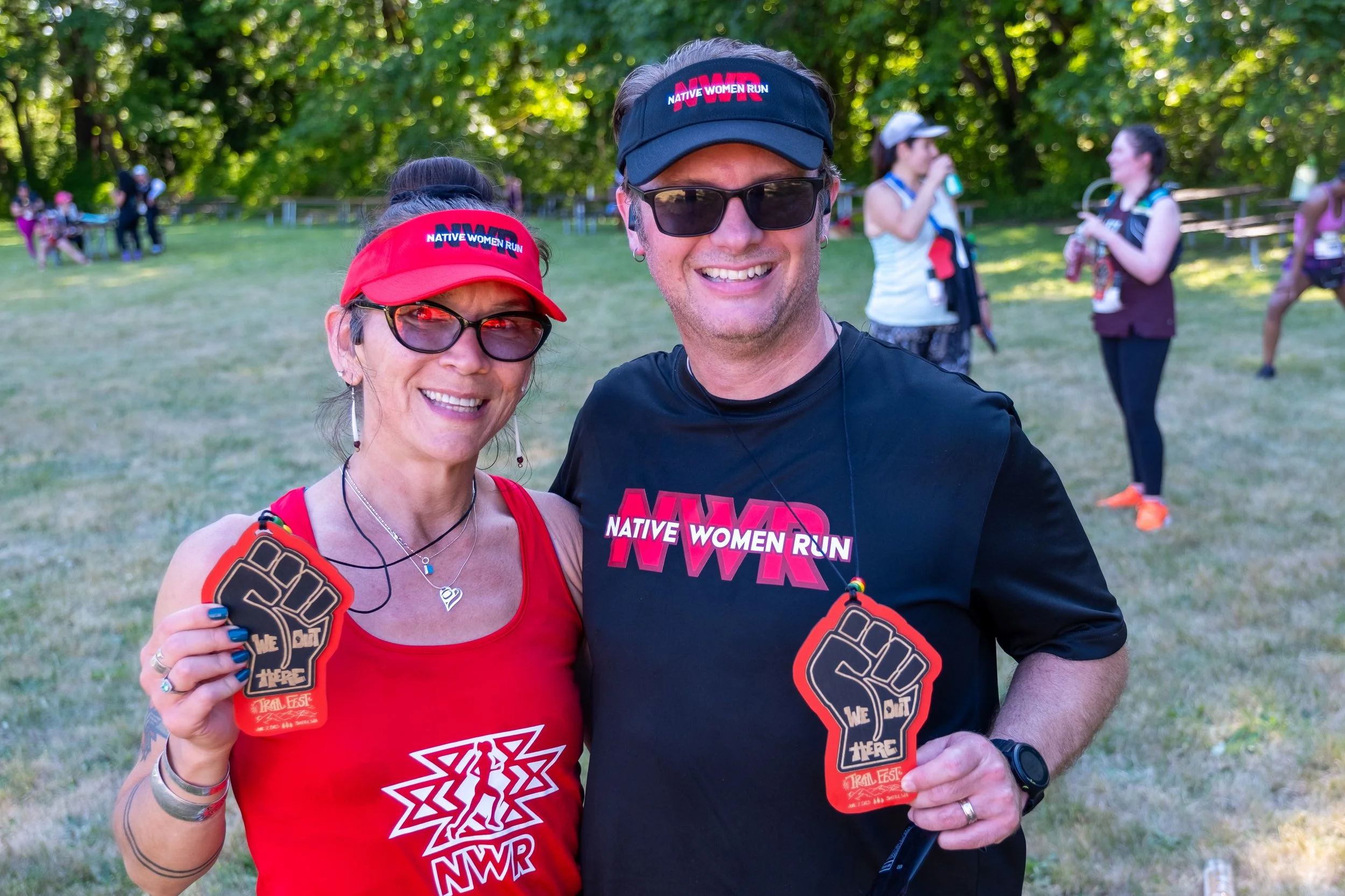Two smiling runners holding medals after a Native Women Run event in a park with other participants in the background.