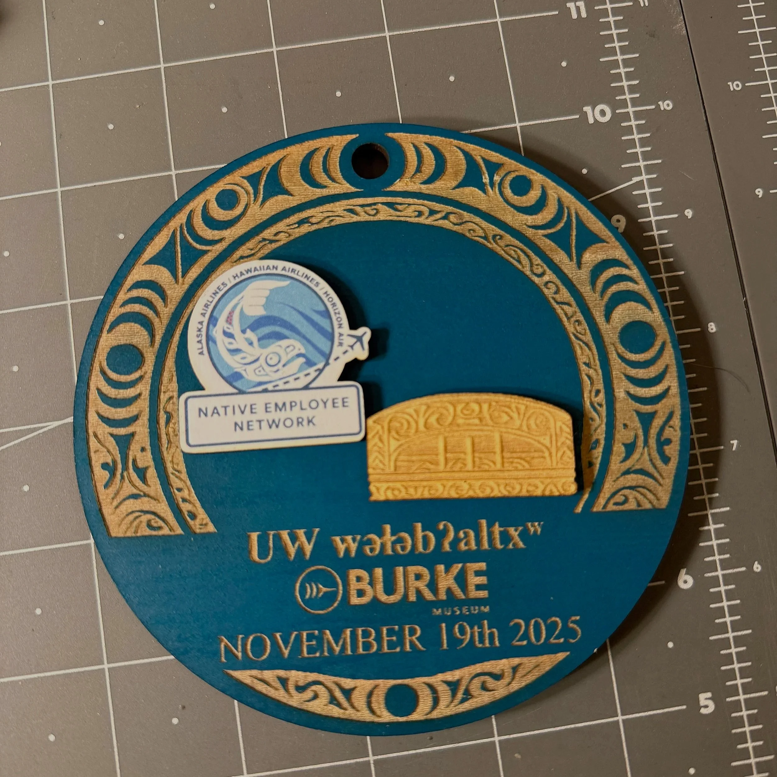 A round badge or coaster with gold and blue colors, featuring a Hawaiian design and the words 'UW Wēkīwāltx™ BURKE MUSEUM' and the date 'November 19th 2025'. It has a logo of a native Hawaiian element and a small rectangular piece resembling a traditional Hawaiian artifact, along with a sticker that says 'Native Employee Network' and a logo of Alaska Airlines, Hawaiian Airlines, and Honu.