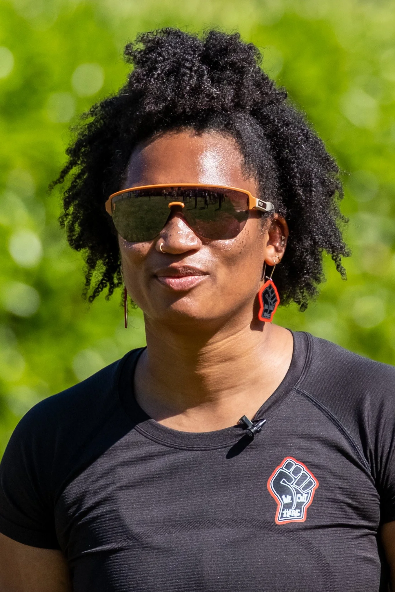 A woman with short curly hair wearing sunglasses, a nose ring, earrings, and a black athletic shirt with a fist logo, outdoors with a green background.
