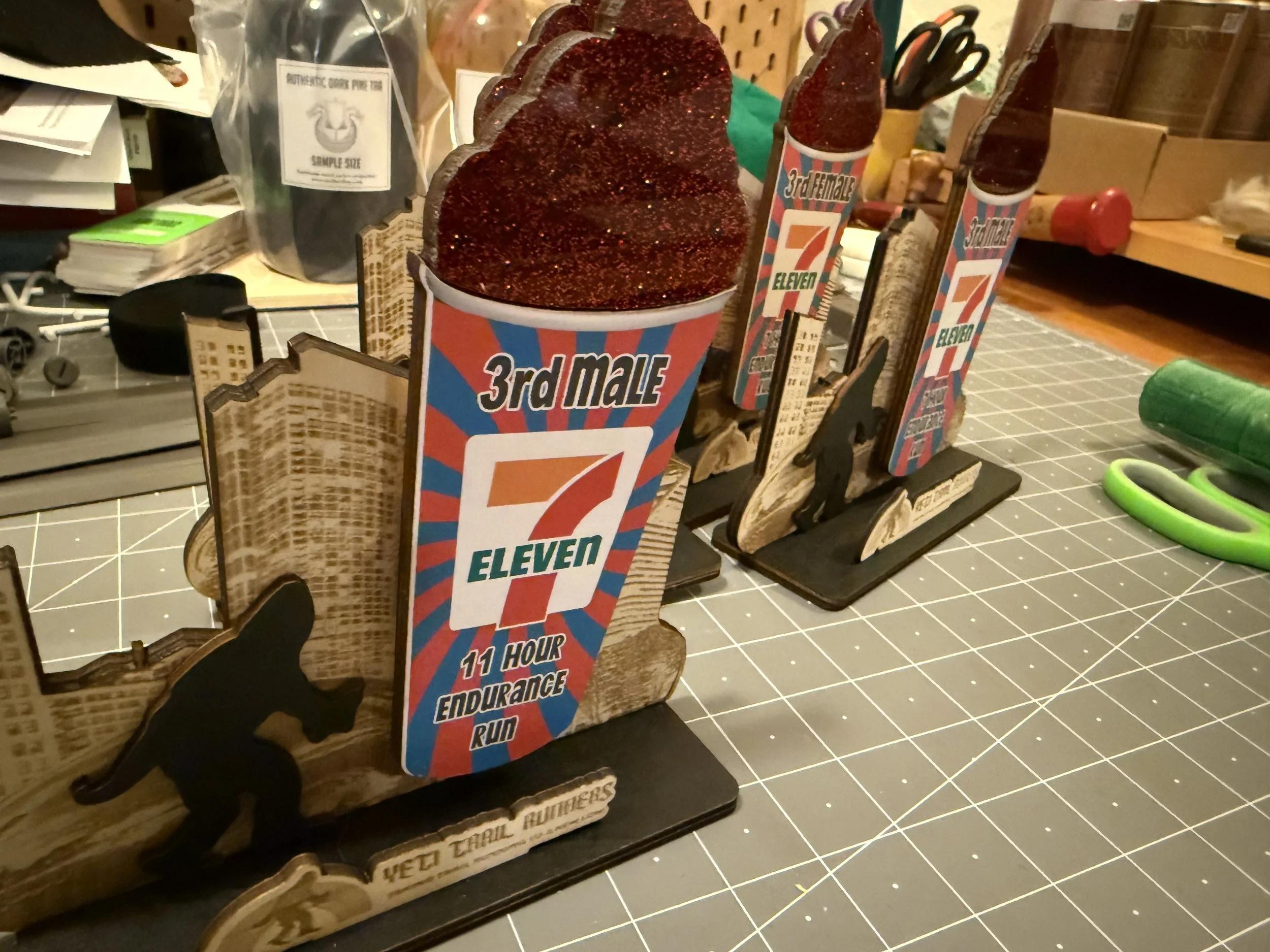 Three wooden trophies with logos of 7-Eleven, featuring a red, white, and blue design, and the text '3rd Male 11 Hour Endurance Run'. The trophies are on a cutting mat with office supplies and small tools in the background.