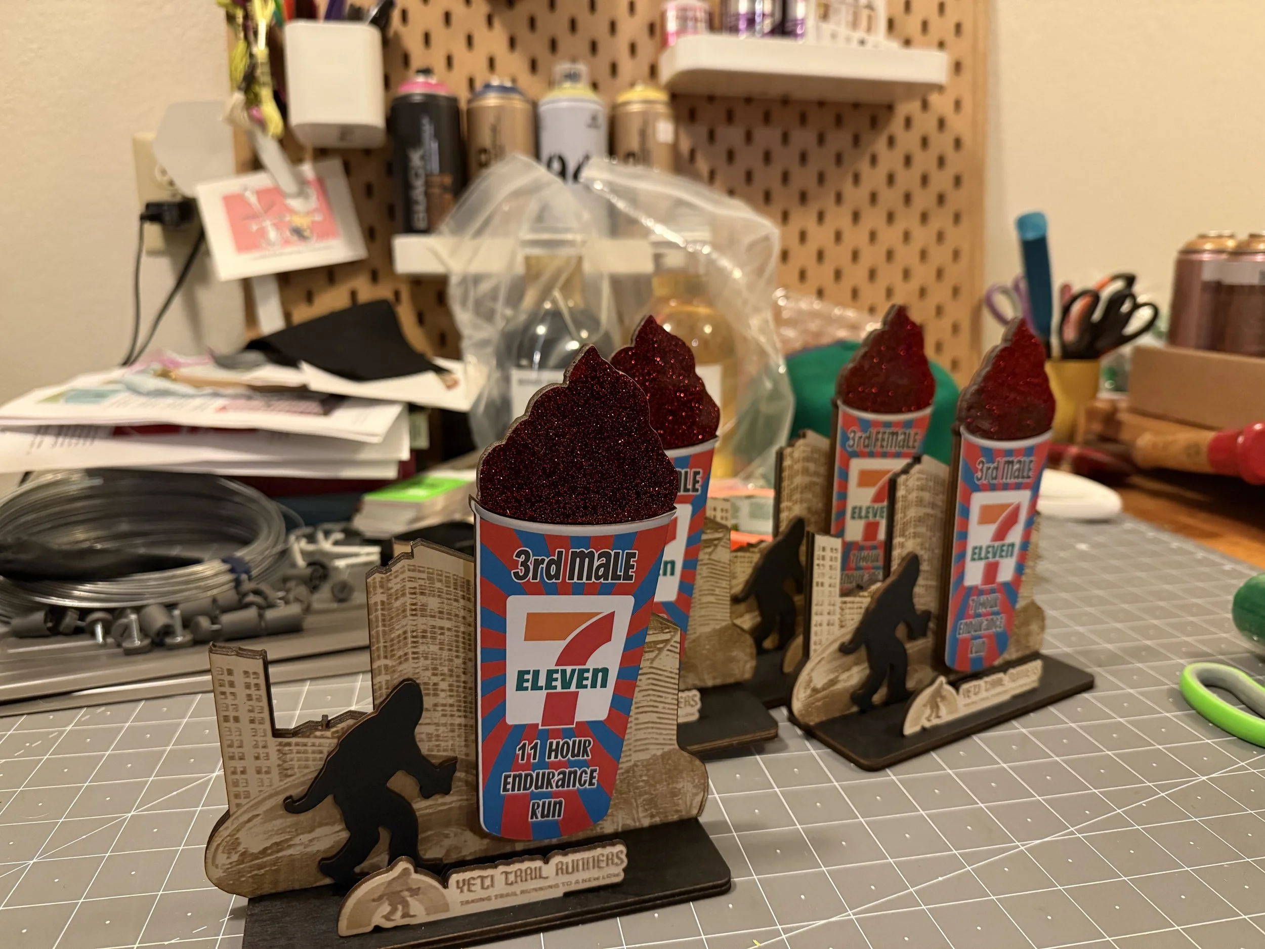 Three souvenir drink cups with red glittery toppers, placed on wooden bases with black silhouettes of mountain climbers for the Yeti Trail Runners Endurance Run, set on a cluttered table with various items in the background.