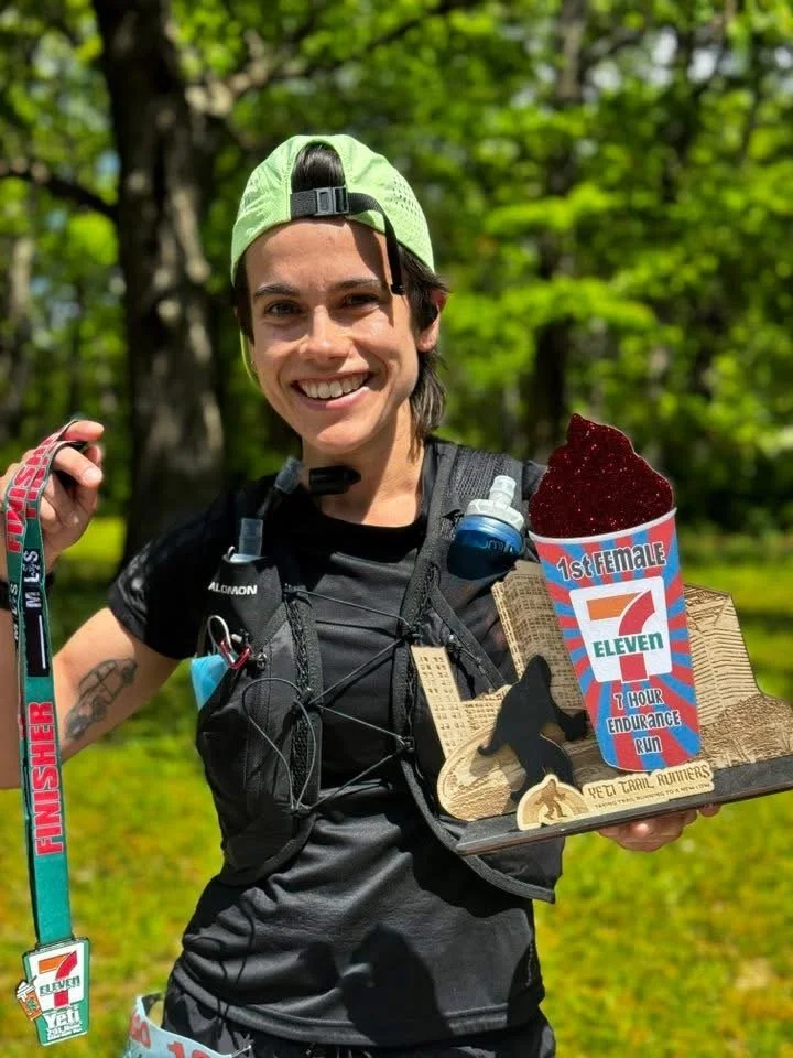 A smiling woman in athletic gear holding a wooden trail award and a colorful container of red ice cream with a label that reads '1st Female 7-Eleven 7 Hour Endurance Run' while wearing a finisher medal around her neck and a backpack, outdoors in a gr