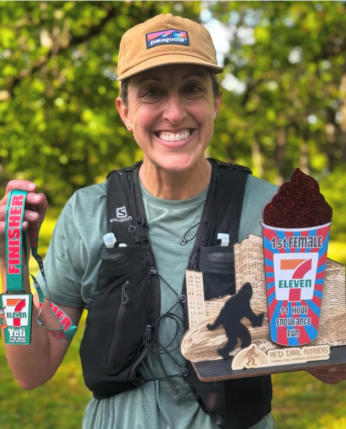 A woman smiling outdoors, wearing a tan Patagonia cap and running gear, holding a finisher medal and a wooden plaque with event symbols, standing among green trees.