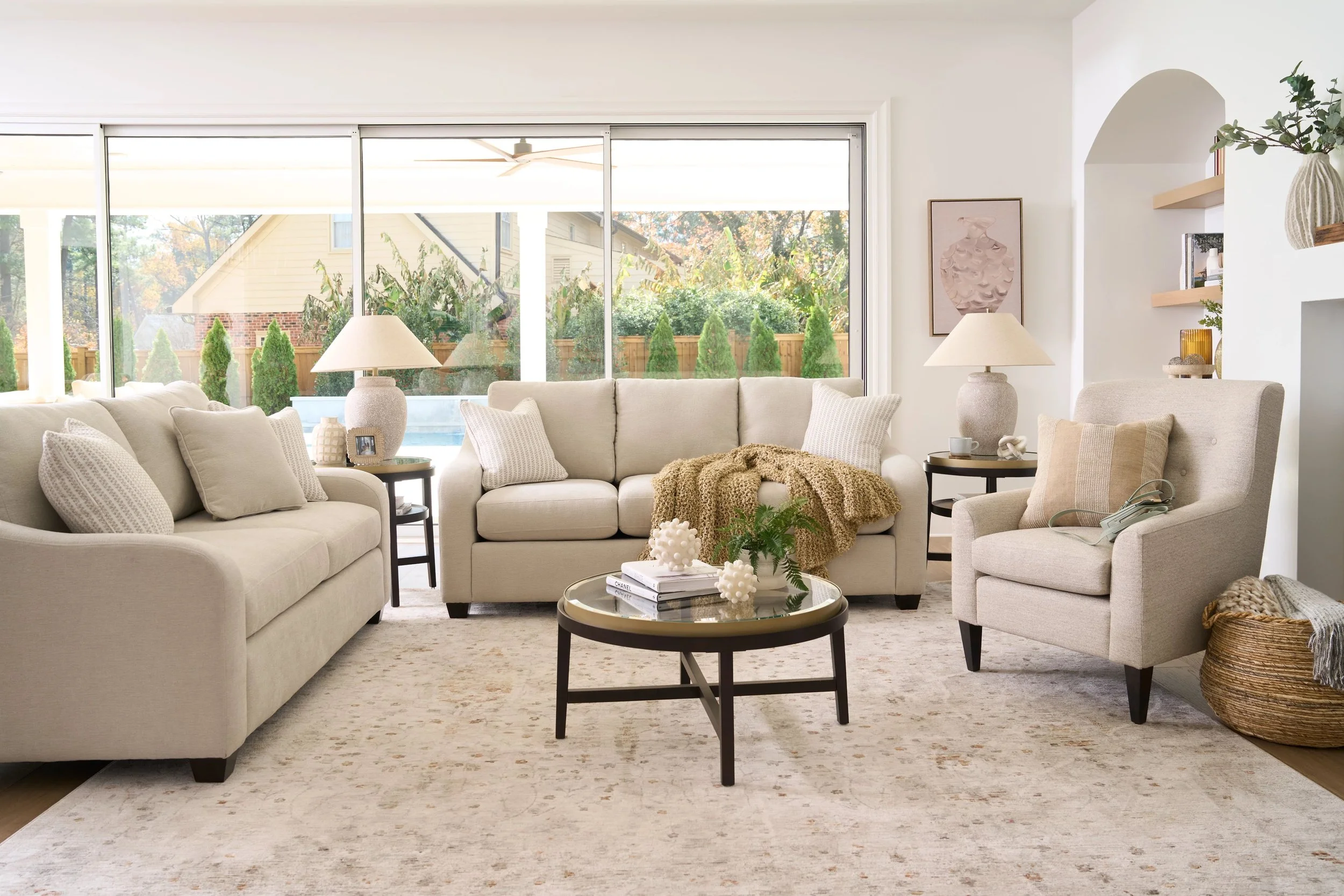 Bright living room with beige sofas and armchair, glass-top coffee table, large windows showing outdoor greenery, and various decorative items like lamps, pillows, and artwork.