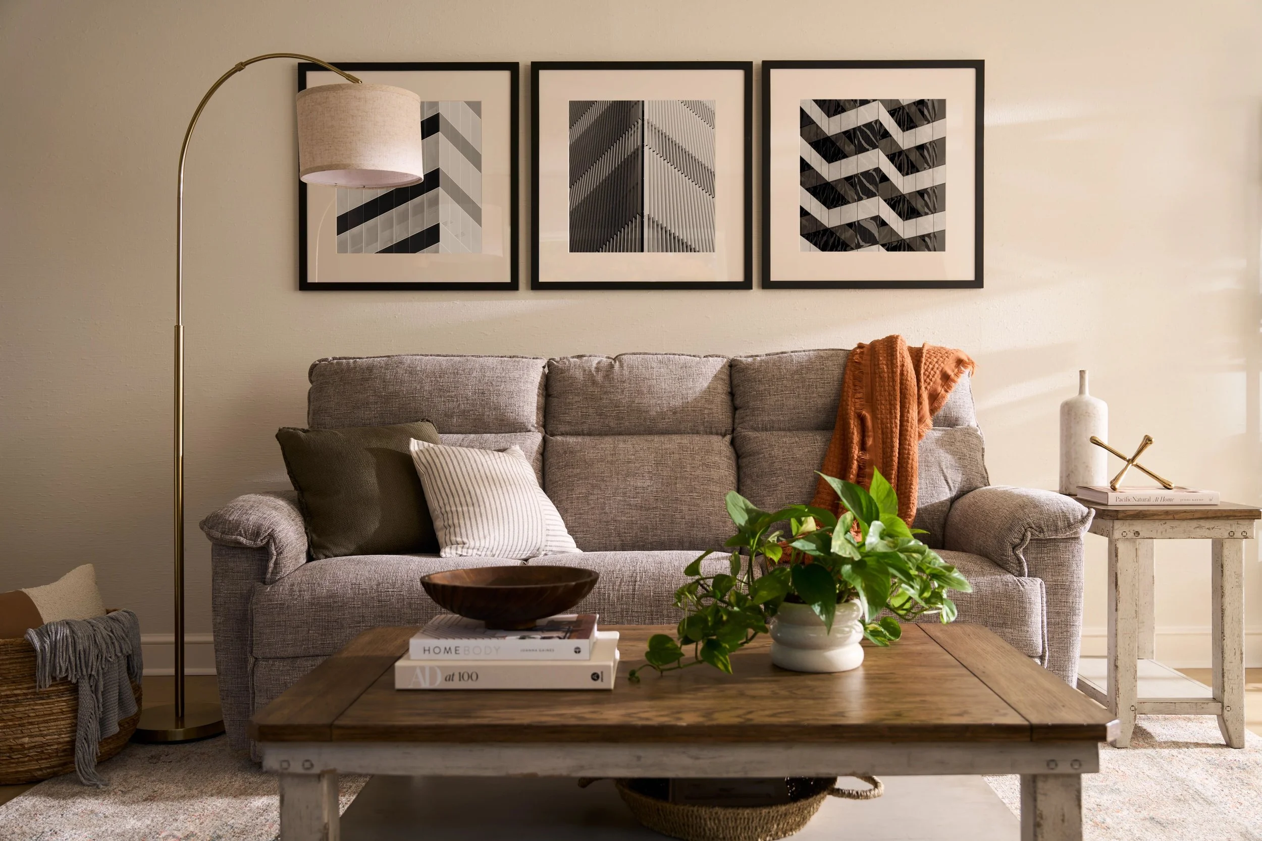 Living room with a gray sofa, framed geometric art above, wooden coffee table with plant and books, white side table with decor, floor lamp, and a basket with a blanket.