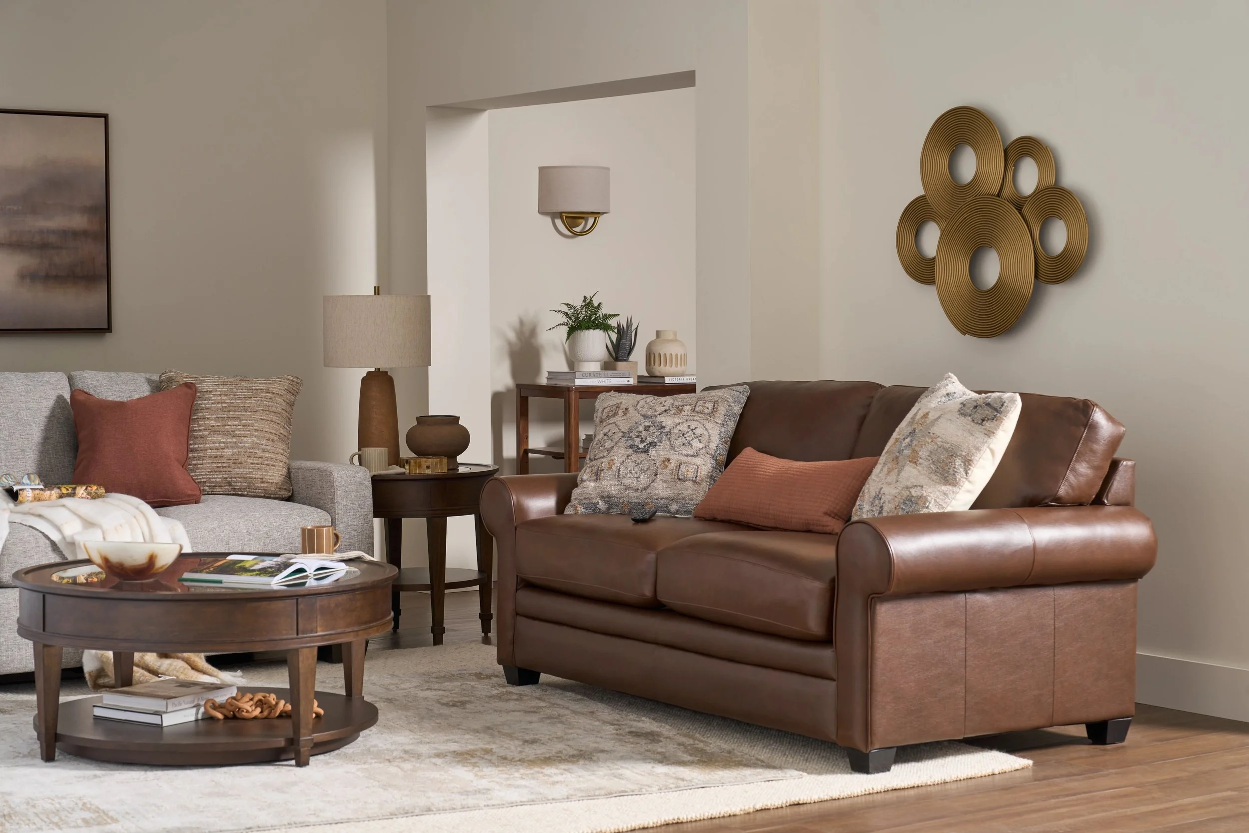 Living room with beige wall, brown leather couch and gray sofa, round wooden coffee table, side table with lamp, decorative wall art, and various pillows.