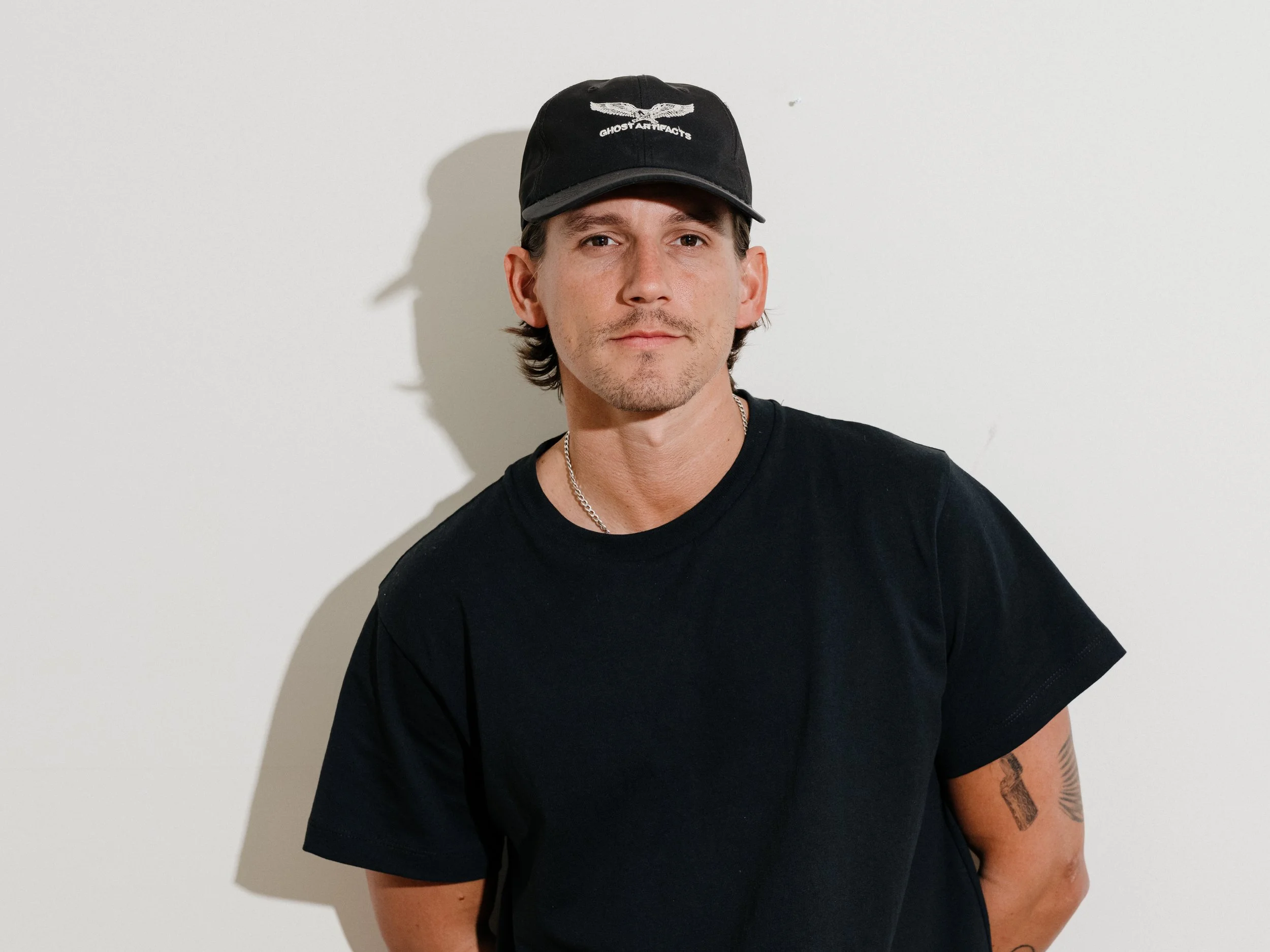 A man standing on the platform of an orange lift, wearing a black satin bomber jacket with patches, indoors near windows with city buildings visible outside.