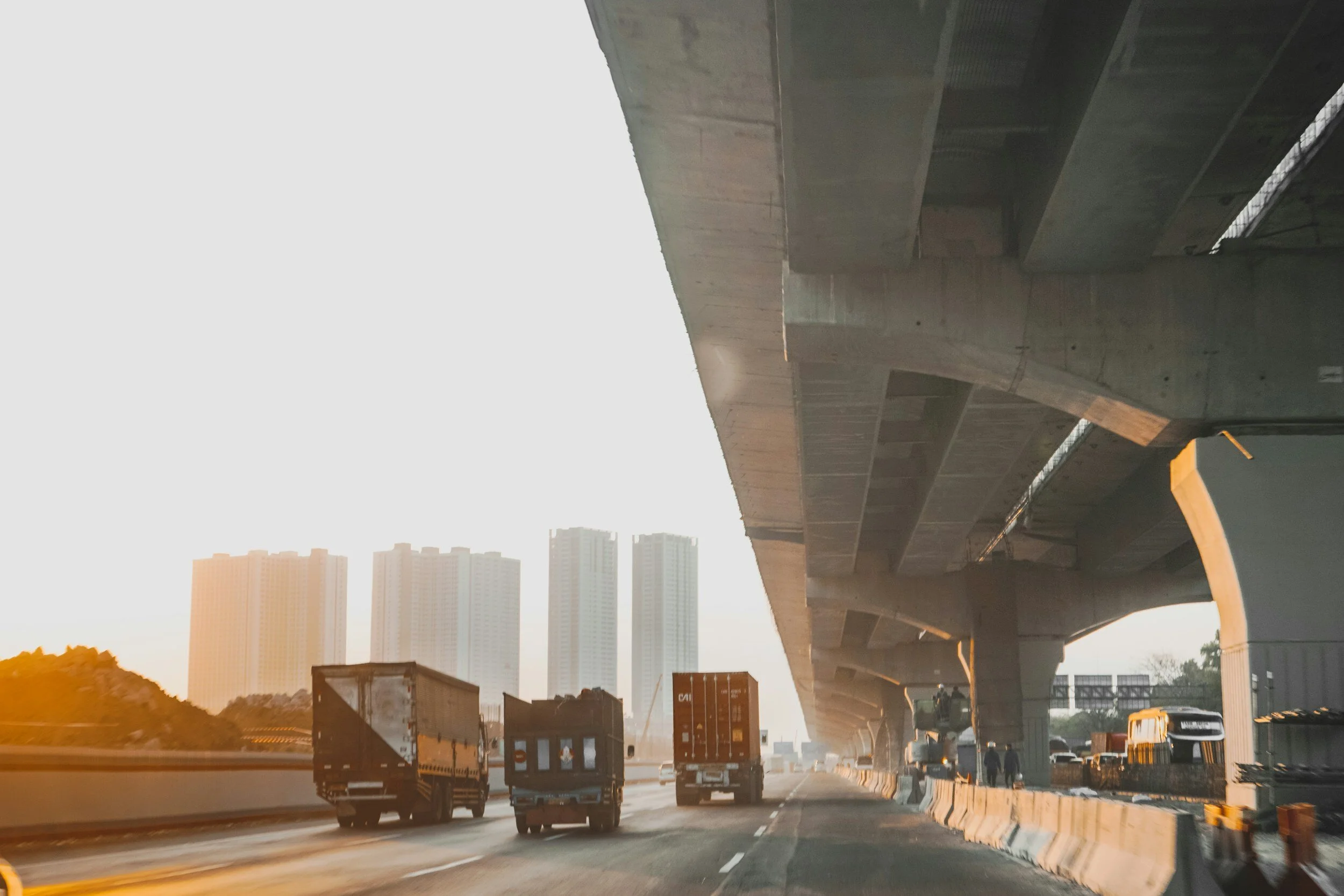 View of a highway with trucks, under an elevated concrete bridge, with tall buildings in the background during sunset