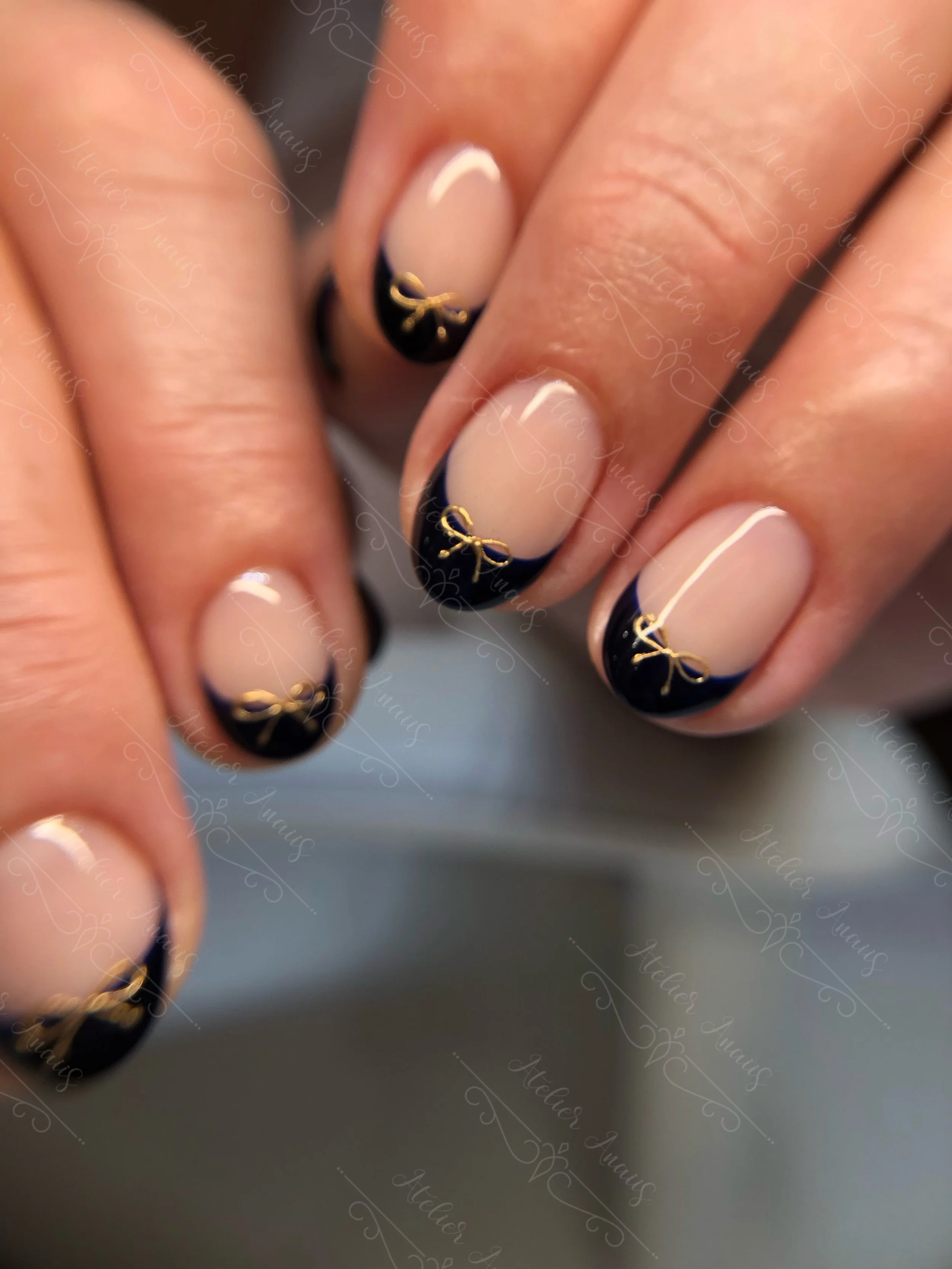 Hands with short natural nails featuring classy black French tip nail art on a glossy nude base with hand drawn Gold Bows.