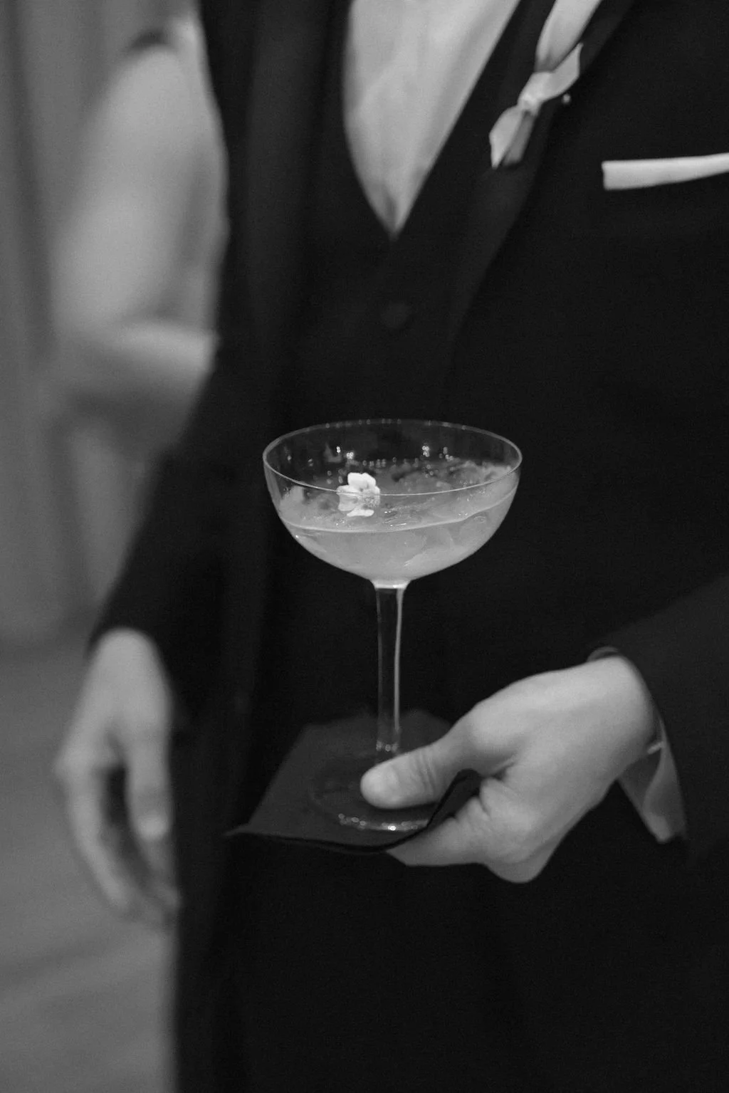 The groom in a black tuxedo holding a cocktail in a coupe glass.