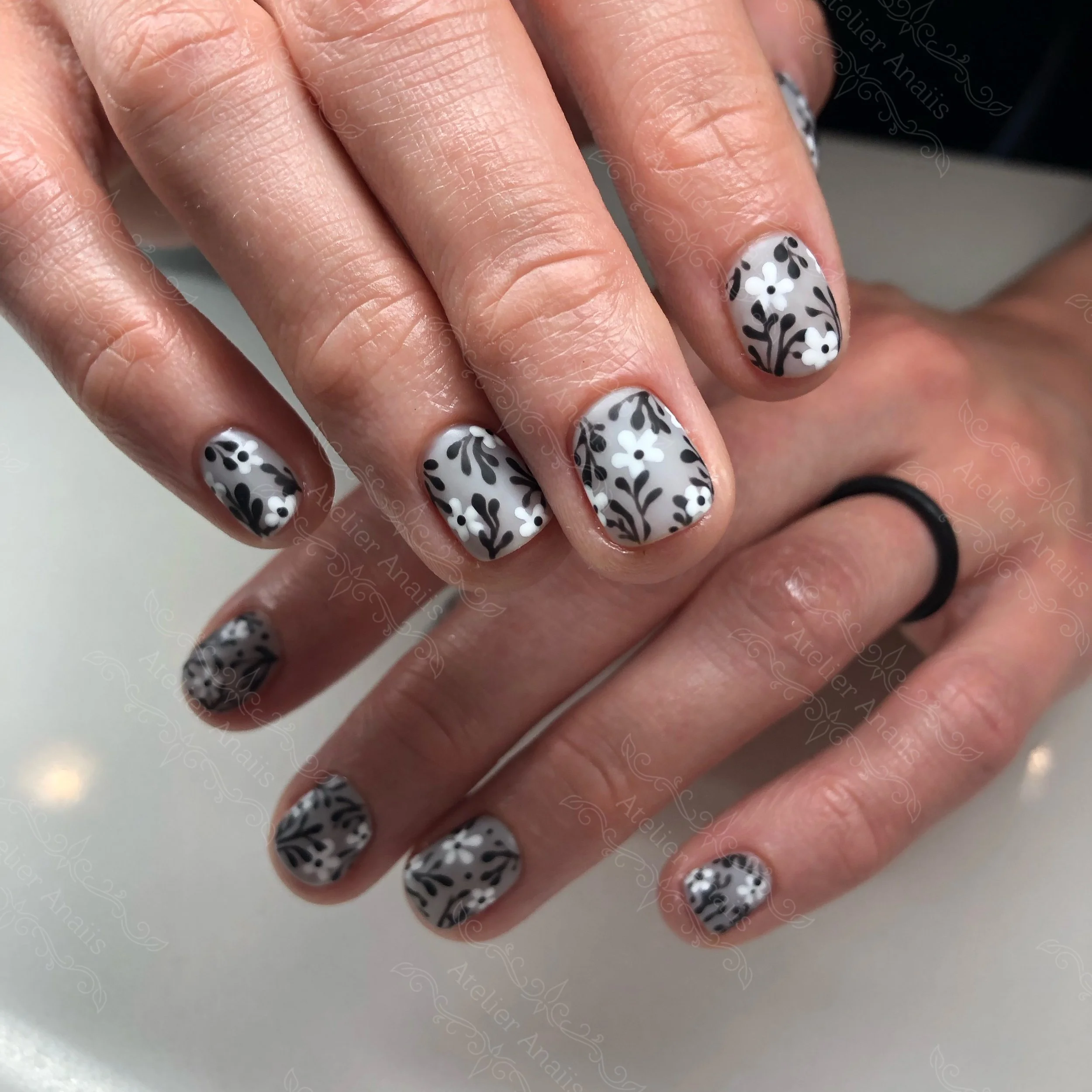 Hands with short natural nails featuring hand drawn black and white flowers nail art on a matte gray base for winter.